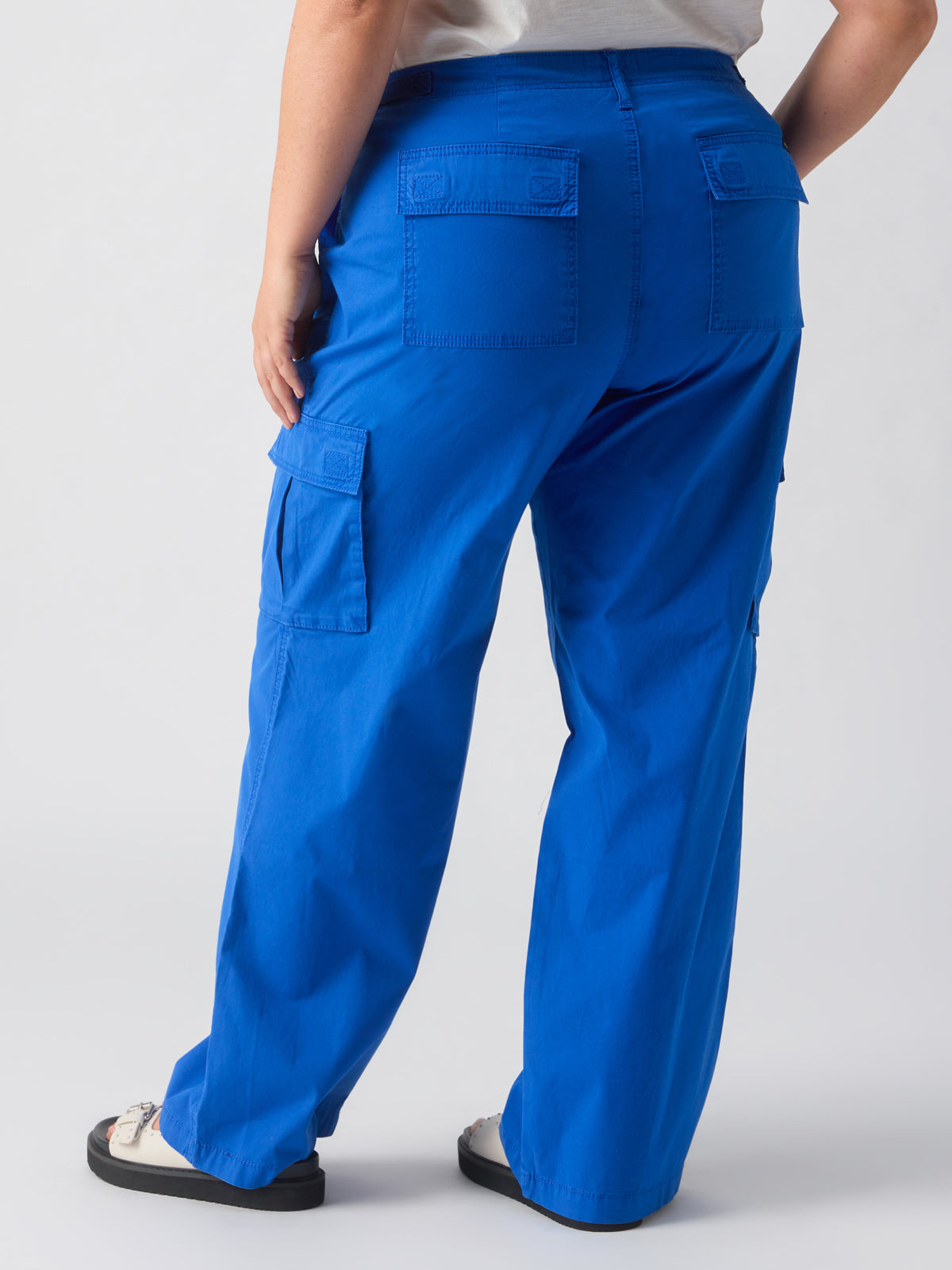 A person is shown from the back, wearing a pair of Sanctuary Clothing's Reissue Cargo Ocean Blue Inclusive Collection pants featuring multiple pockets and a white shirt. The person is also wearing sandals paired with white socks against a plain, light-colored background.