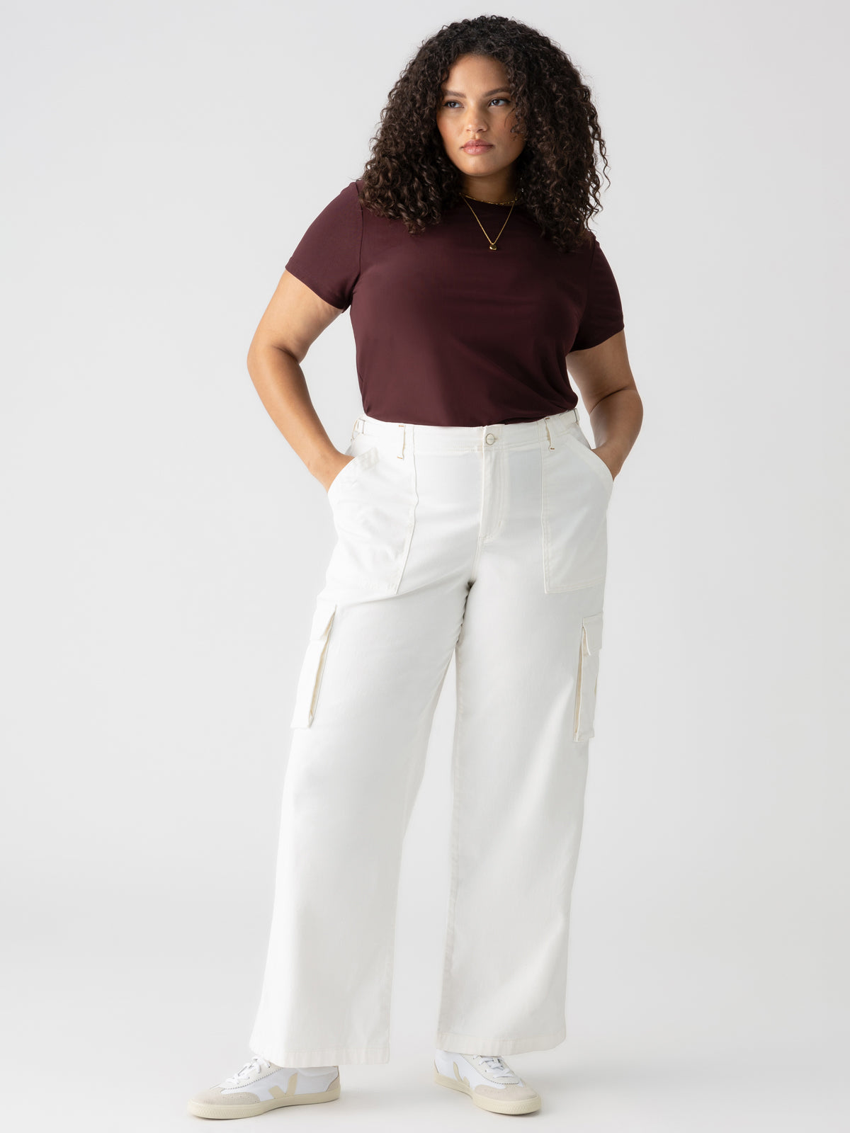 A person with curly hair stands against a plain background, hands in pockets, wearing a maroon short-sleeved top and the Sanctuary Clothing Reissue Cargo White Inclusive Collection pants with side cargo pockets, accessorized with a necklace and white sneakers.