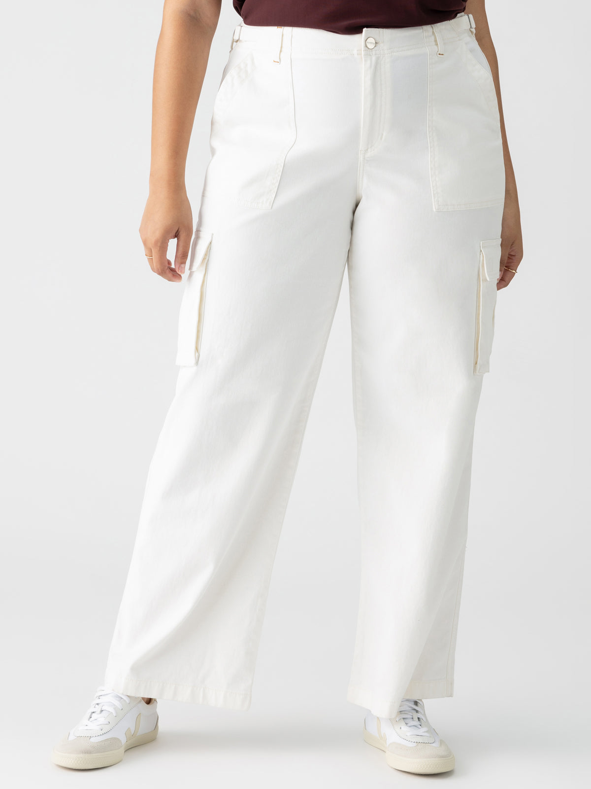 A person is wearing the Reissue Cargo White Inclusive Collection pants by Sanctuary Clothing. The high-waisted, wide-leg cargo pants with side pockets are paired with white sneakers. The person's upper outfit and head remain outside the frame, against a plain white background.