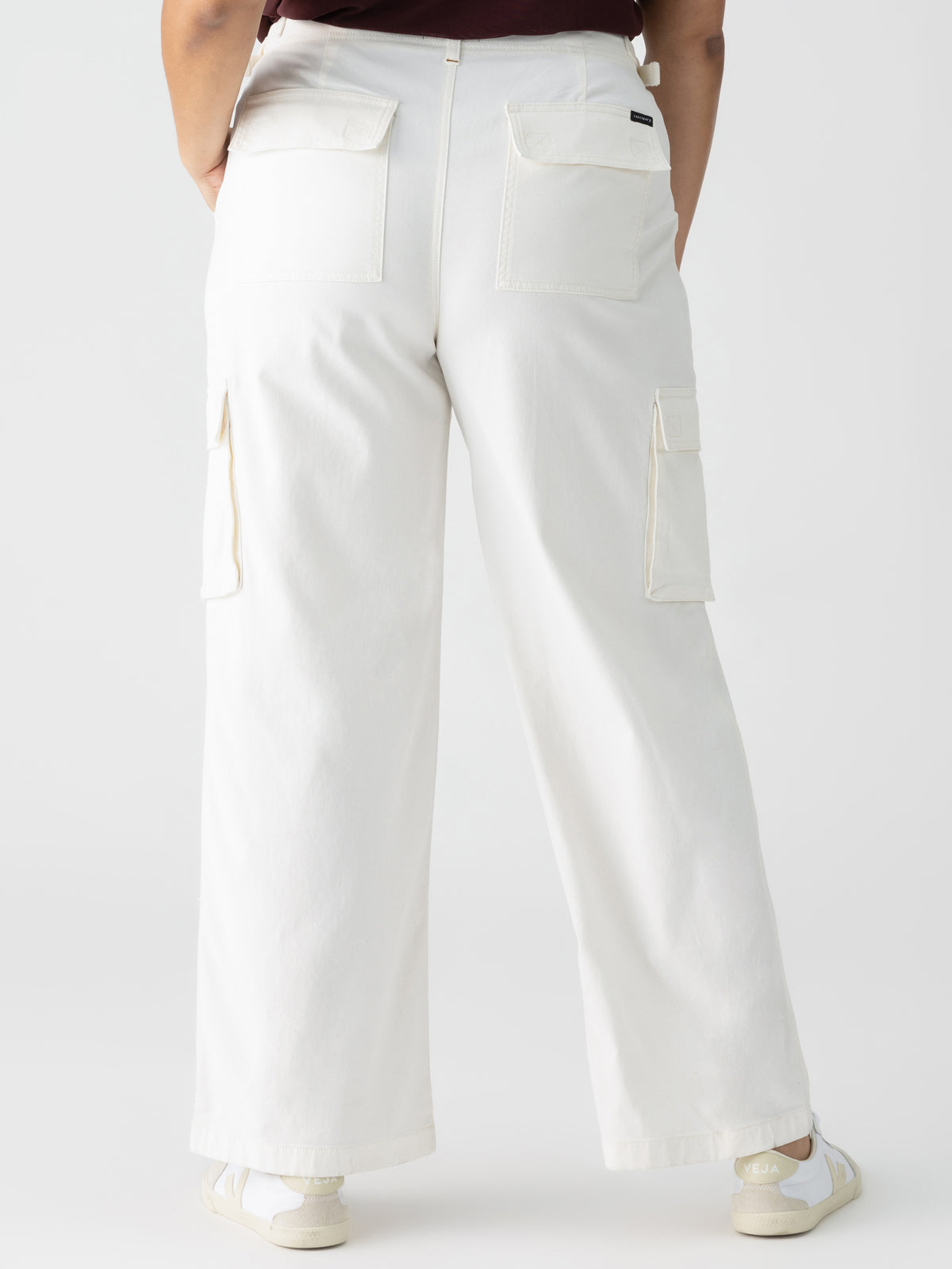 A person wearing Sanctuary Clothing's Reissue Cargo White Inclusive Collection wide-leg cargo pants, featuring multiple pockets including two large rear pockets and side cargo pockets, stands with their back facing the camera. They also wear a dark top and white sneakers. The background is plain and light-colored.