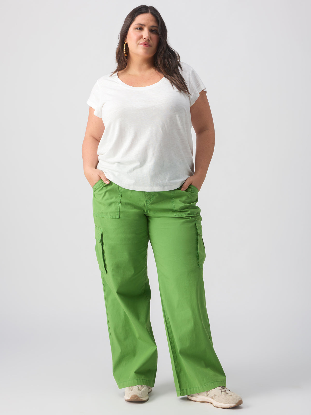 A woman stands confidently against a plain background, wearing a white short-sleeved top, Sanctuary Clothing's Reissue Cargo Washed Chlorophyl Inclusive Collection bright green cargo pants with side pockets, and white sneakers. She has her hands in her pockets and wears gold hoop earrings.