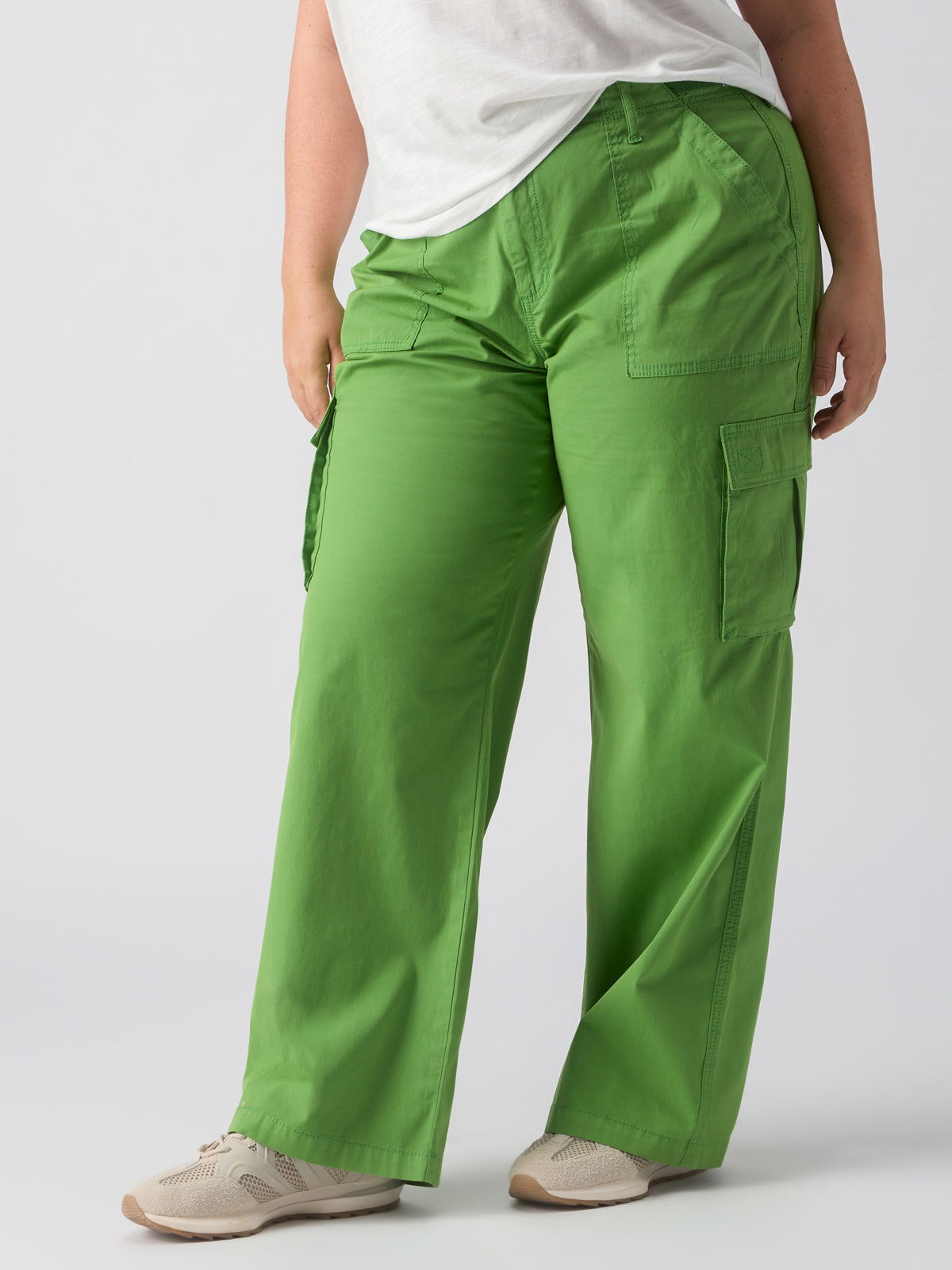 A person is wearing the Reissue Cargo Washed Chlorophyl Inclusive Collection pants from Sanctuary Clothing, paired with white sneakers. The person's upper body is partially cropped out of the shot, while the background showcases a plain light gray color.