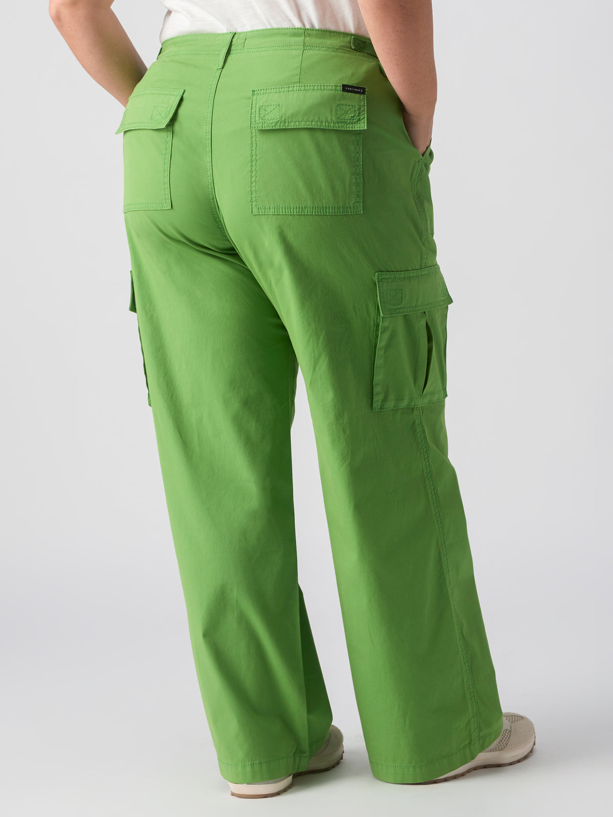 A person wearing bright green cargo pants from Sanctuary Clothing's Reissue Cargo Washed Chlorophyl Inclusive Collection, which feature multiple pockets on the back and sides, stands with their hands in their pockets. The person is seen from behind and is also wearing a white shirt and beige shoes.