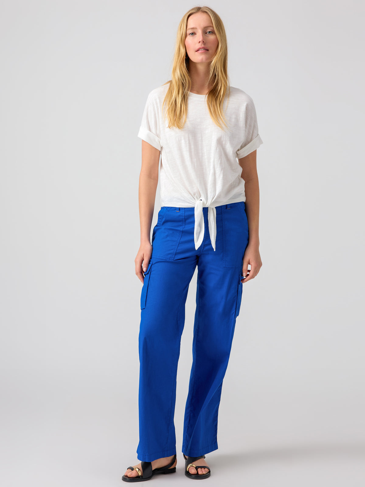 A person with long blonde hair is wearing a white short-sleeve top tied in a knot at the waist and the Reissue Cargo Ocean Blue pants from Sanctuary Clothing. They are paired with black sandals. The background is a plain, light color.