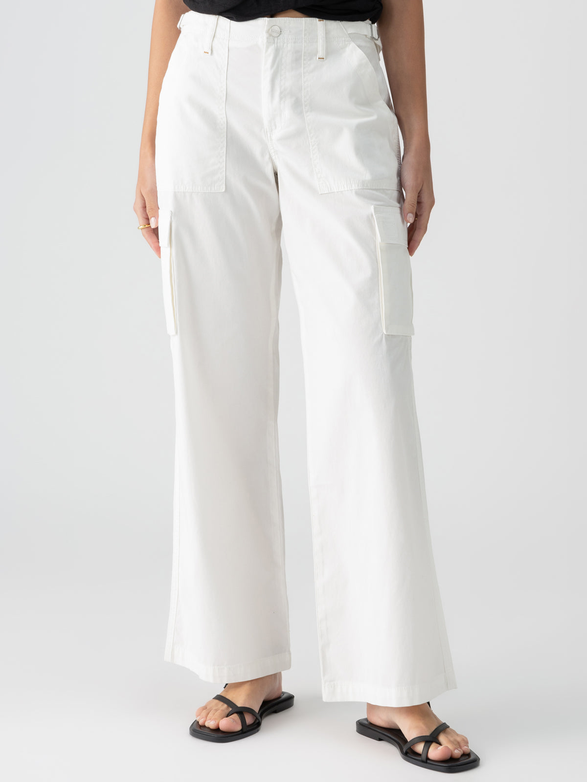 A person is dressed in Sanctuary Clothing's Reissue Cargo White pants, featuring high-waisted, wide-leg design and large thigh pockets with hands partially tucked in. They are matched with black strappy sandals and a black top peeking out from the bottom. The background is plain white.