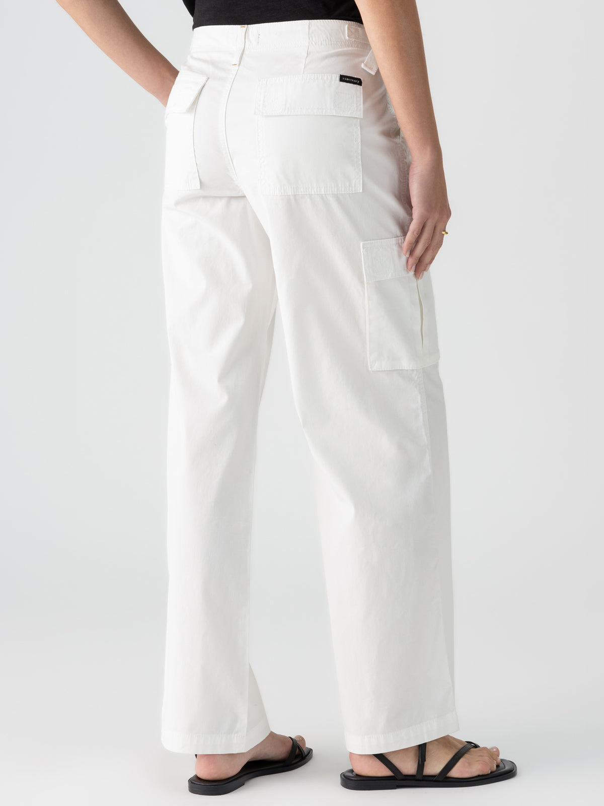 A person dressed in a black top and Sanctuary Clothing’s Reissue Cargo White high-waisted utility pants with multiple pockets stands facing away. They are wearing black sandals and have a hand resting on their hip. The background is plain white.