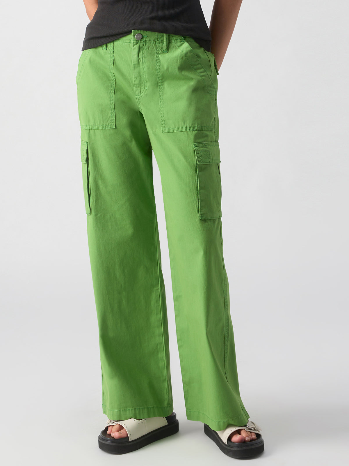 A person is wearing Sanctuary Clothing's Reissue Cargo Washed Chlorophyl, which are bright green cargo pants that feature side pockets and a button closure. They are also sporting black sandals with white soles. The image highlights the lower half of the individual against a plain white background.