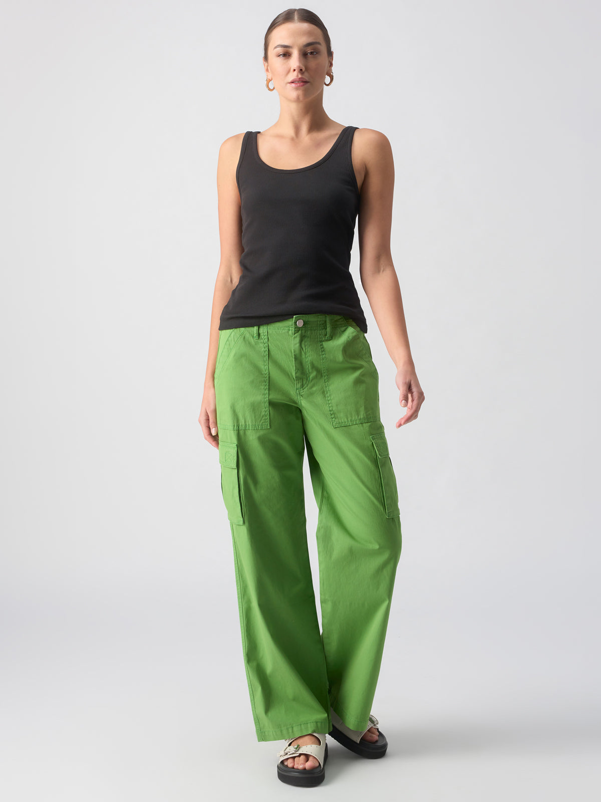 A person with tied-back hair is wearing a black tank top, Sanctuary Clothing's Reissue Cargo Washed Chlorophyll pants, and black sandals. They are standing against a plain white background, looking straight ahead with one arm by their side and the other slightly bent.