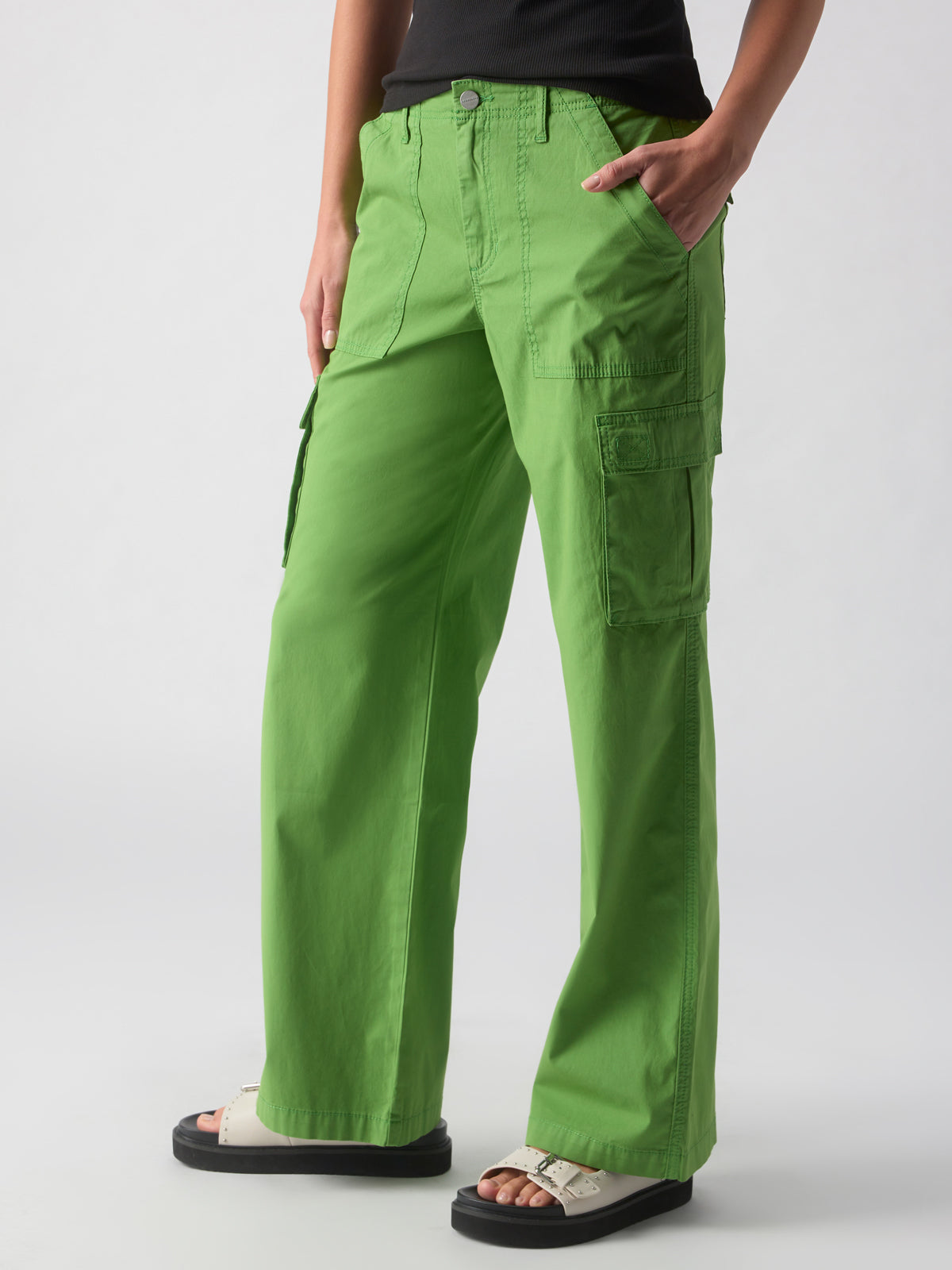 A person is shown wearing the Reissue Cargo Washed Chlorophyl pants from Sanctuary Clothing, paired with white sandals. The vibrant green pants feature multiple pockets, adding both functionality and style. The person is standing with one hand in a pocket against a plain, light-colored background.