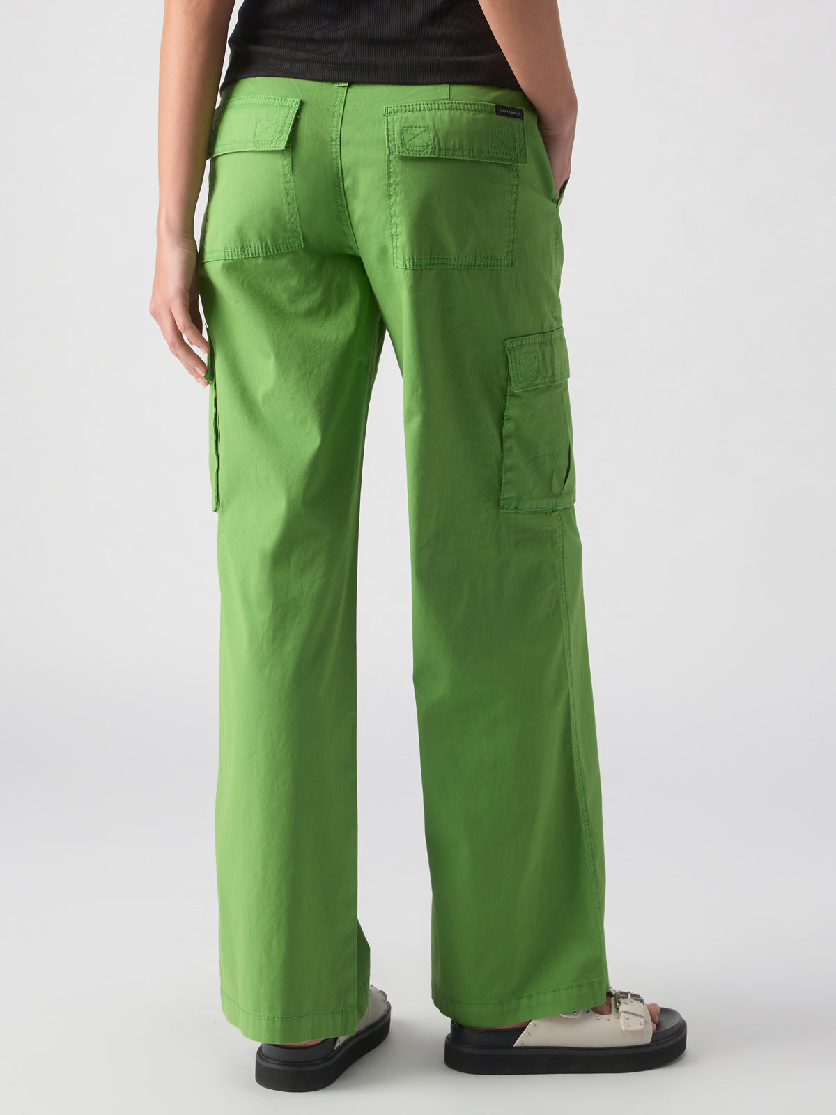 A person wearing the Reissue Cargo Washed Chlorophyl pants from Sanctuary Clothing and white sneakers is shown from the back, highlighting the pants' multiple pockets and relaxed fit. The plain, neutral background emphasizes the vibrant color and design of these bright green cargo pants.