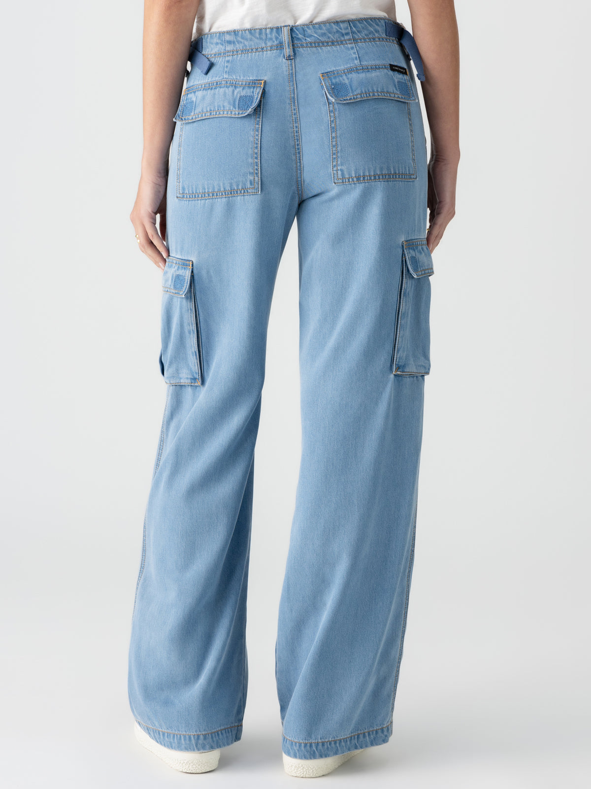 A person wearing the Reissue Cargo Pant Sun Drenched by Sanctuary Clothing stands with their back to the camera. The light blue, high-waisted cargo jeans feature large back pockets with flaps and side cargo pockets. The person pairs this with a white top and white sneakers.