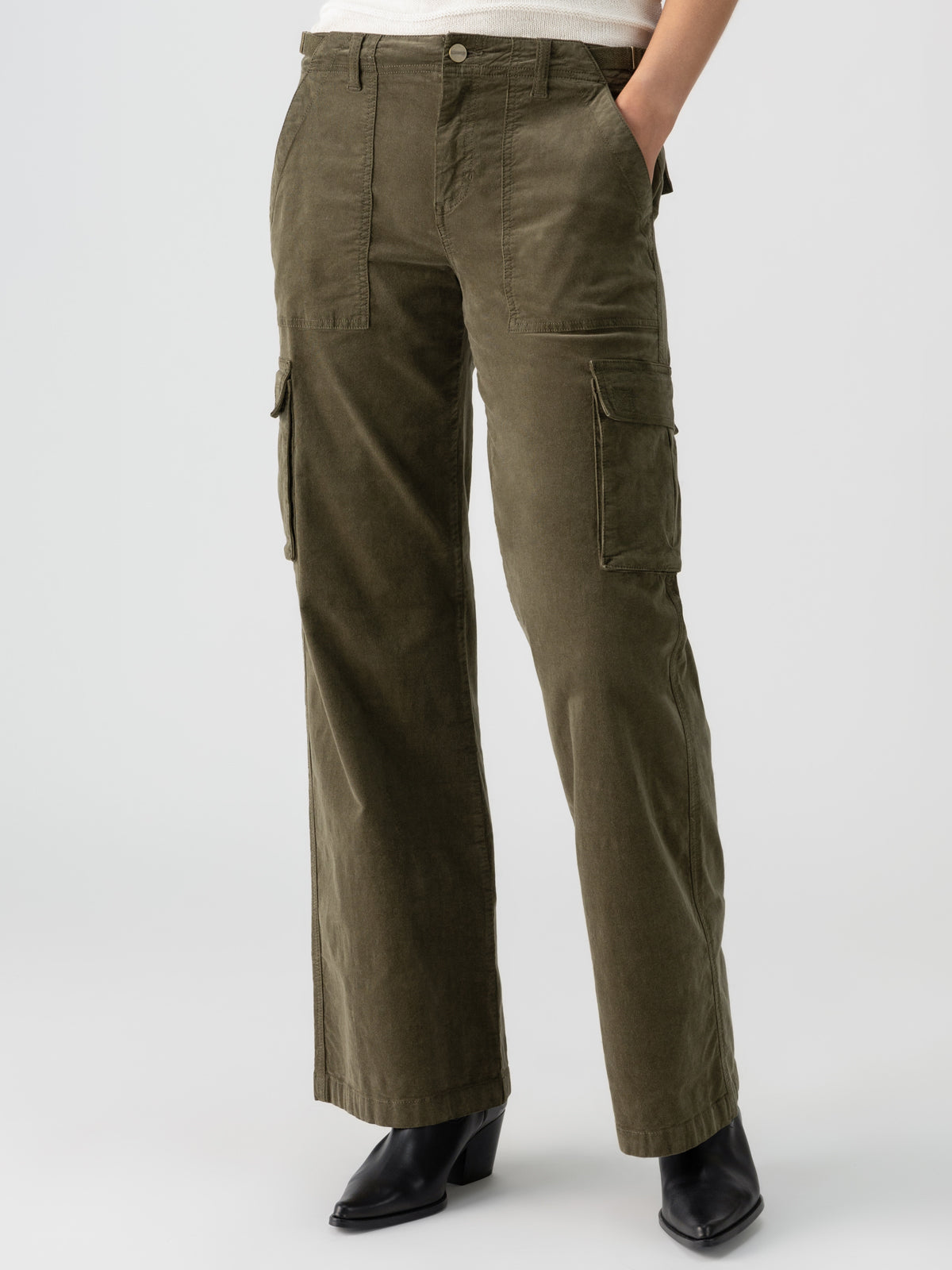 A model is showcasing the Reissue Cargo Burnt Olive pants from Sanctuary Clothing, featuring a high-waisted design and large thigh pockets. The model pairs these olive green cargo pants with black ankle boots and a light-colored top that is partially tucked in. The background is plain white, highlighting the outfit.