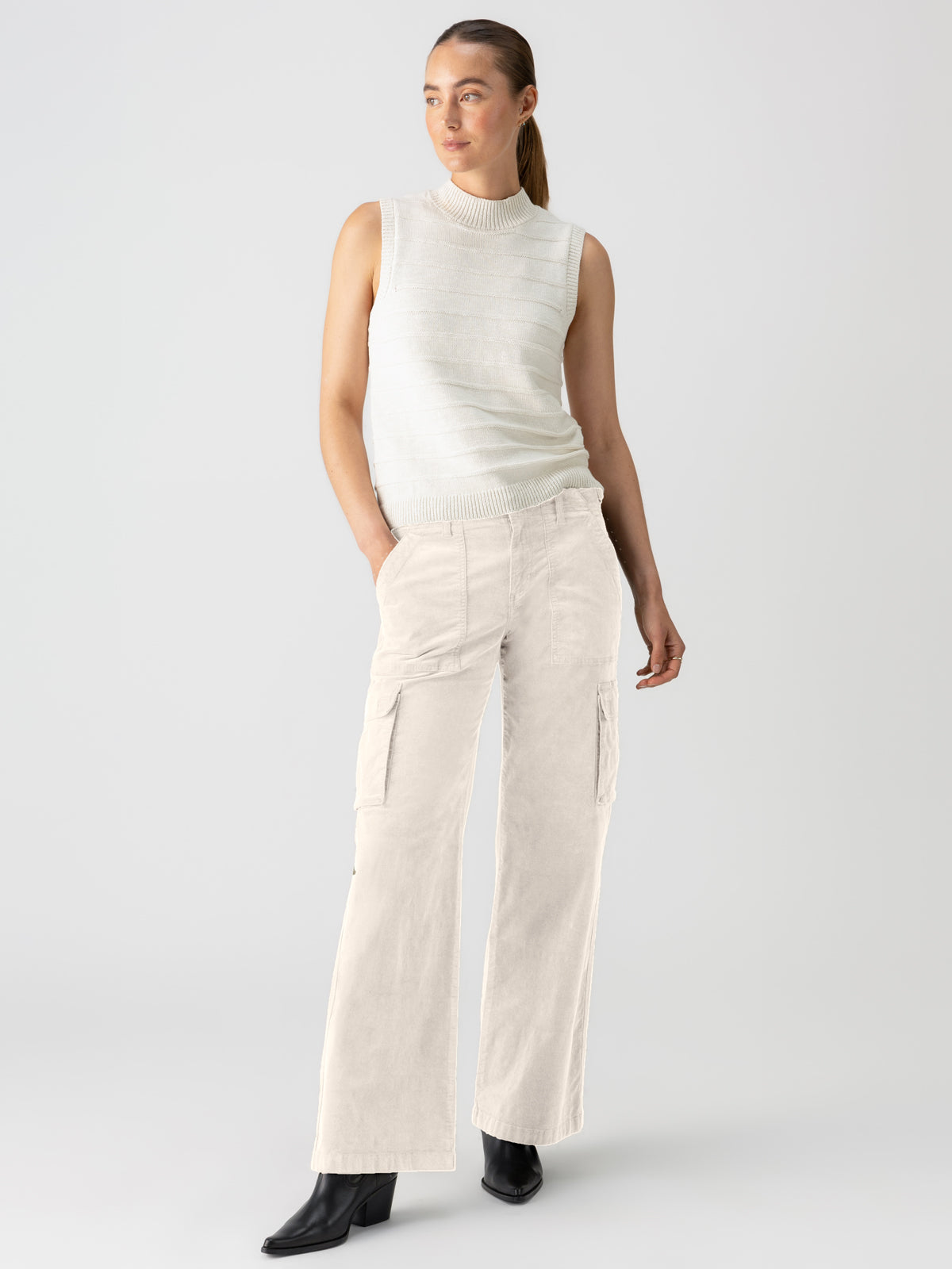 A person with long hair tied back is wearing a sleeveless, light-colored knit top and the high-waisted, wide-leg Reissue Cargo Toasted Almond pants from Sanctuary Clothing. They have their hands in the pants' pockets and are standing against a plain, light background, looking slightly to the side.