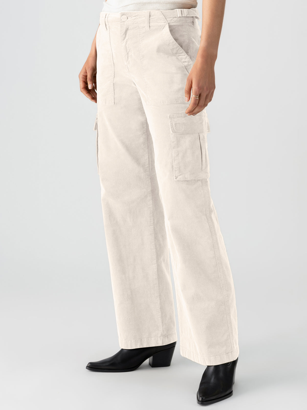 A person is shown from the waist down wearing the Reissue Cargo Toasted Almond from Sanctuary Clothing, paired with black heeled ankle boots. The off-white, high-waisted cargo pants feature multiple pockets, including two large side pockets. The background is neutral and light gray.