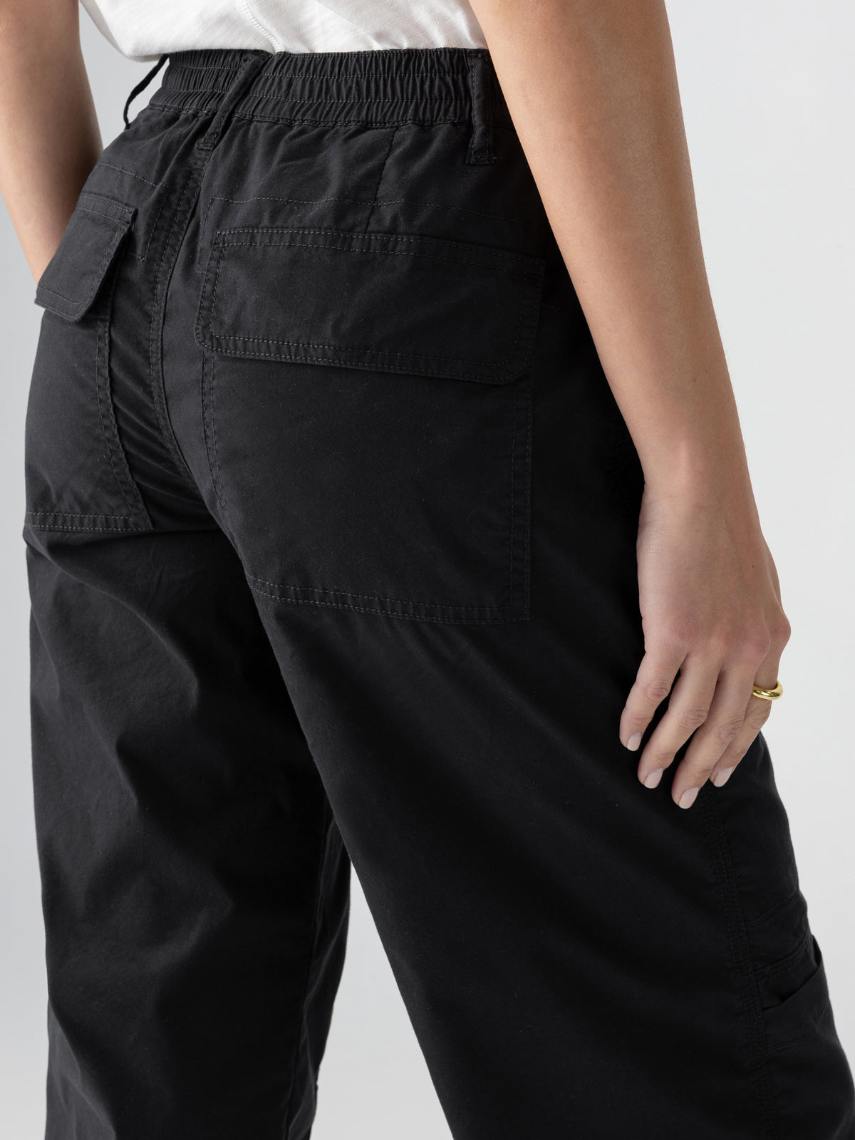 A person in a white top is shown from the back, wearing Sanctuary Clothing's Cali Cargo Pant Black, which features large back pockets and an elastic waistband. The individual is wearing a gold ring on their right hand, which rests next to the side pocket. The background is a plain light gray.