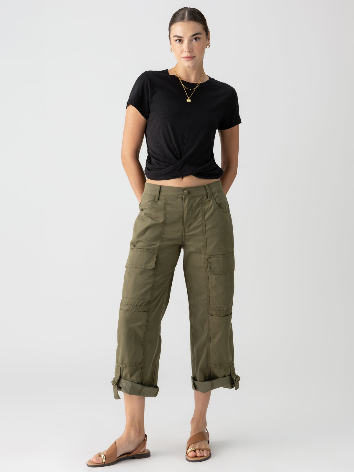 A person stands against a plain background, wearing a black knotted t-shirt, Sanctuary Clothing's Cali Cargo Pant in Mossy Green with rolled-up cuffs, and brown sandals. They have their hair tied back and are accessorized with a gold necklace and small earrings.