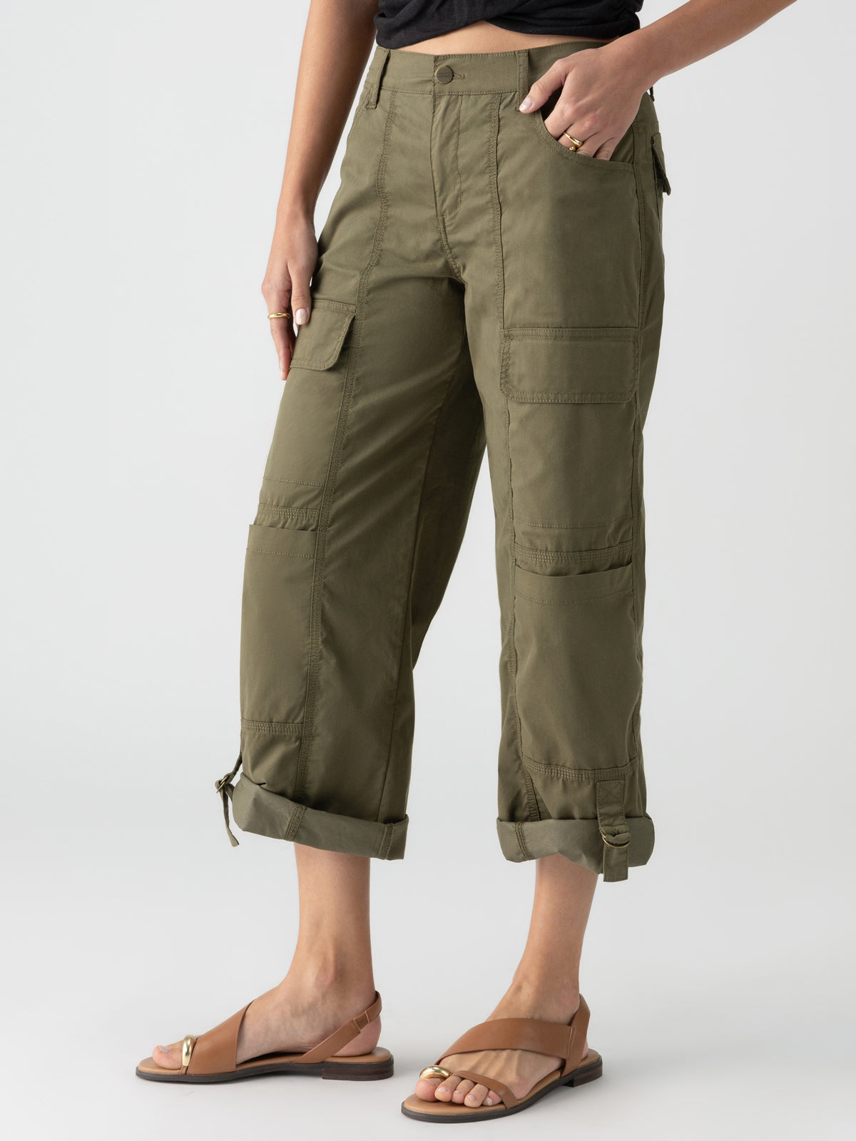 Person wearing Sanctuary Clothing's Cali Cargo Pant in Mossy Green, which features multiple pockets and rolled-up cuffs, paired with tan flat sandals. The person’s hand is resting in their pocket, and they are standing against a plain background. Only the lower half of the body is visible.