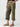 Person wearing Sanctuary Clothing's Cali Cargo Pant in Mossy Green, which features multiple pockets and rolled-up cuffs, paired with tan flat sandals. The person’s hand is resting in their pocket, and they are standing against a plain background. Only the lower half of the body is visible.