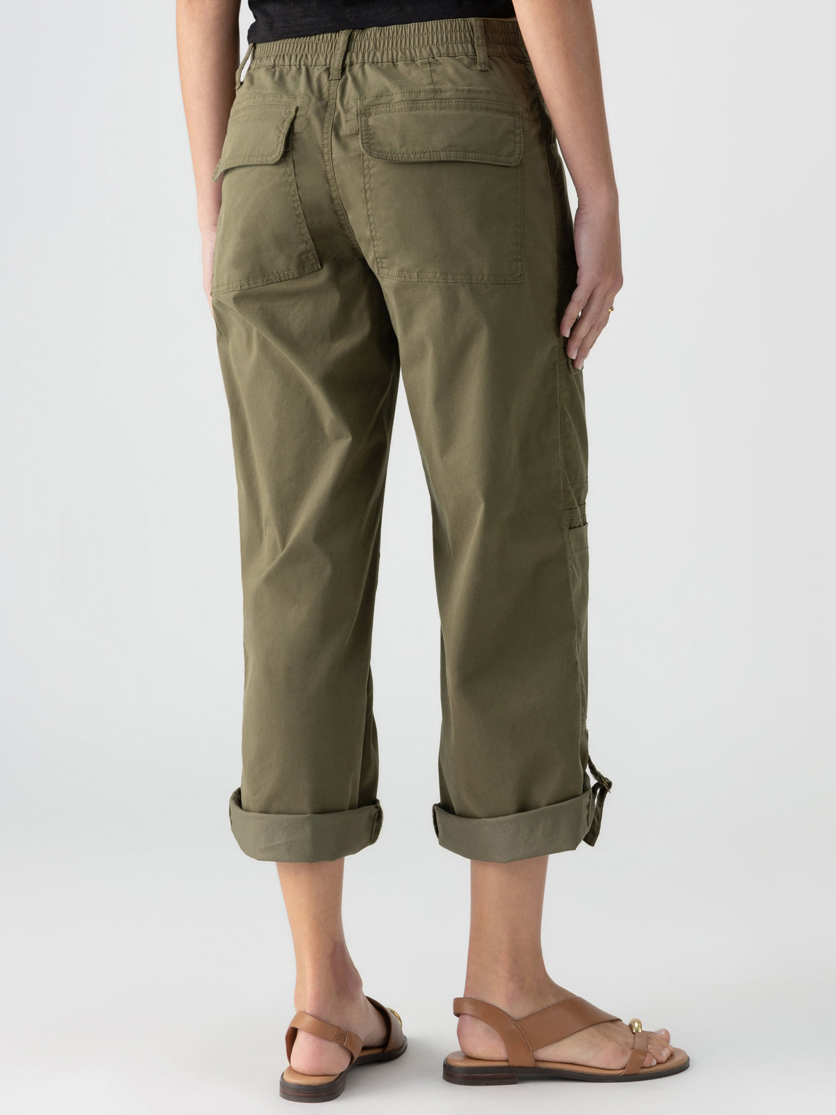 A person is standing with their back to the camera, wearing the Cali Cargo Pant in Mossy Green by Sanctuary Clothing, featuring rolled-up cuffs and cargo pockets on the back. They have paired it with a black shirt and brown sandals with ankle straps. The background is plain and light-colored.