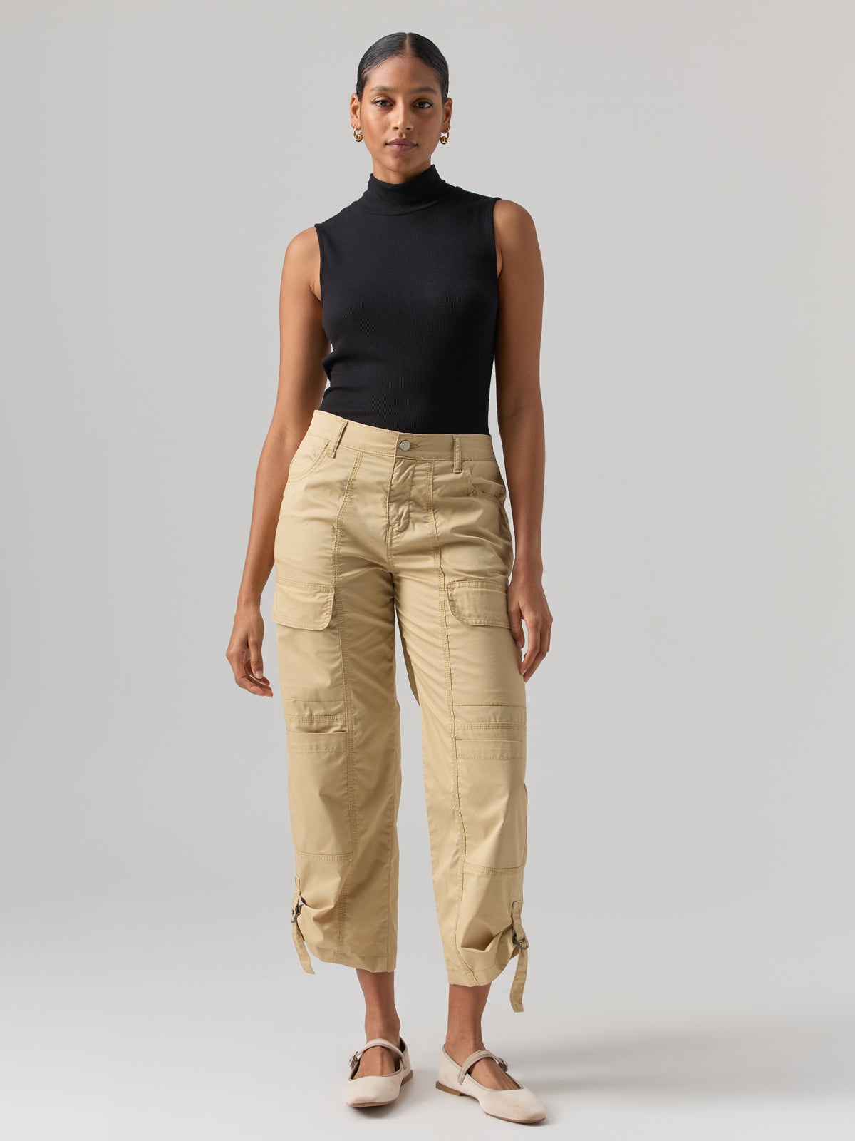 A woman stands facing forward against a plain background. She is wearing a sleeveless black turtleneck top, Sanctuary Clothing's Cali Cargo Pant in True Khaki, featuring multiple pockets and cinched hems, and light-colored flat shoes. Her hair is pulled back, and she has a neutral expression.