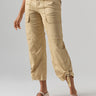 A person is standing against a plain background, modeling the Sanctuary Clothing Cali Cargo Pant in True Khaki. The pants feature multiple pockets and zippered cuffs. The person is also wearing light-colored flats, with their hands relaxed by their sides.