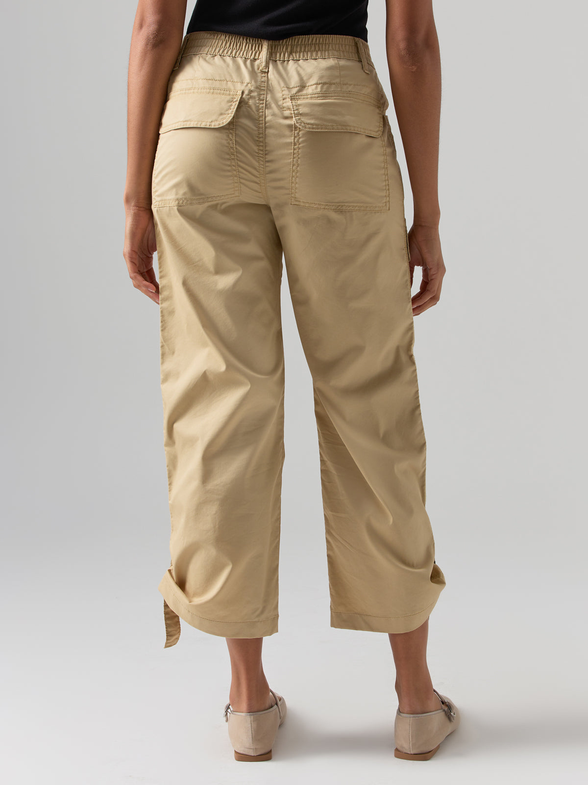 A person is standing with their back to the camera, wearing the Cali Cargo Pant True Khaki from Sanctuary Clothing, which are high-waisted cargo pants in beige with two large back pockets. They are also wearing light beige shoes and a black top. The background is plain and light-colored.