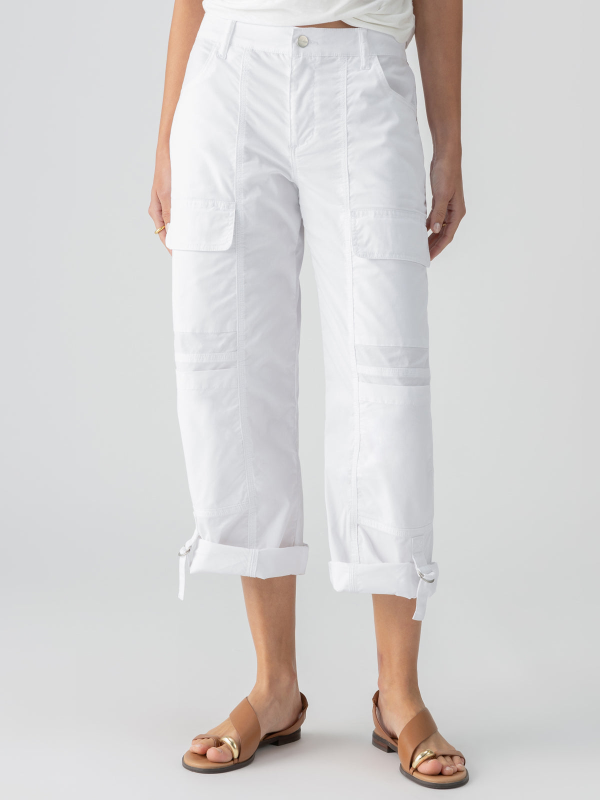 A person is wearing the Sanctuary Clothing's Cali Cargo Pant in white, featuring multiple pockets and drawstring cuffs, paired with a white tucked-in shirt. They are also sporting brown open-toe sandals. The photo captures the individual from the shoulders down, standing against a plain background.