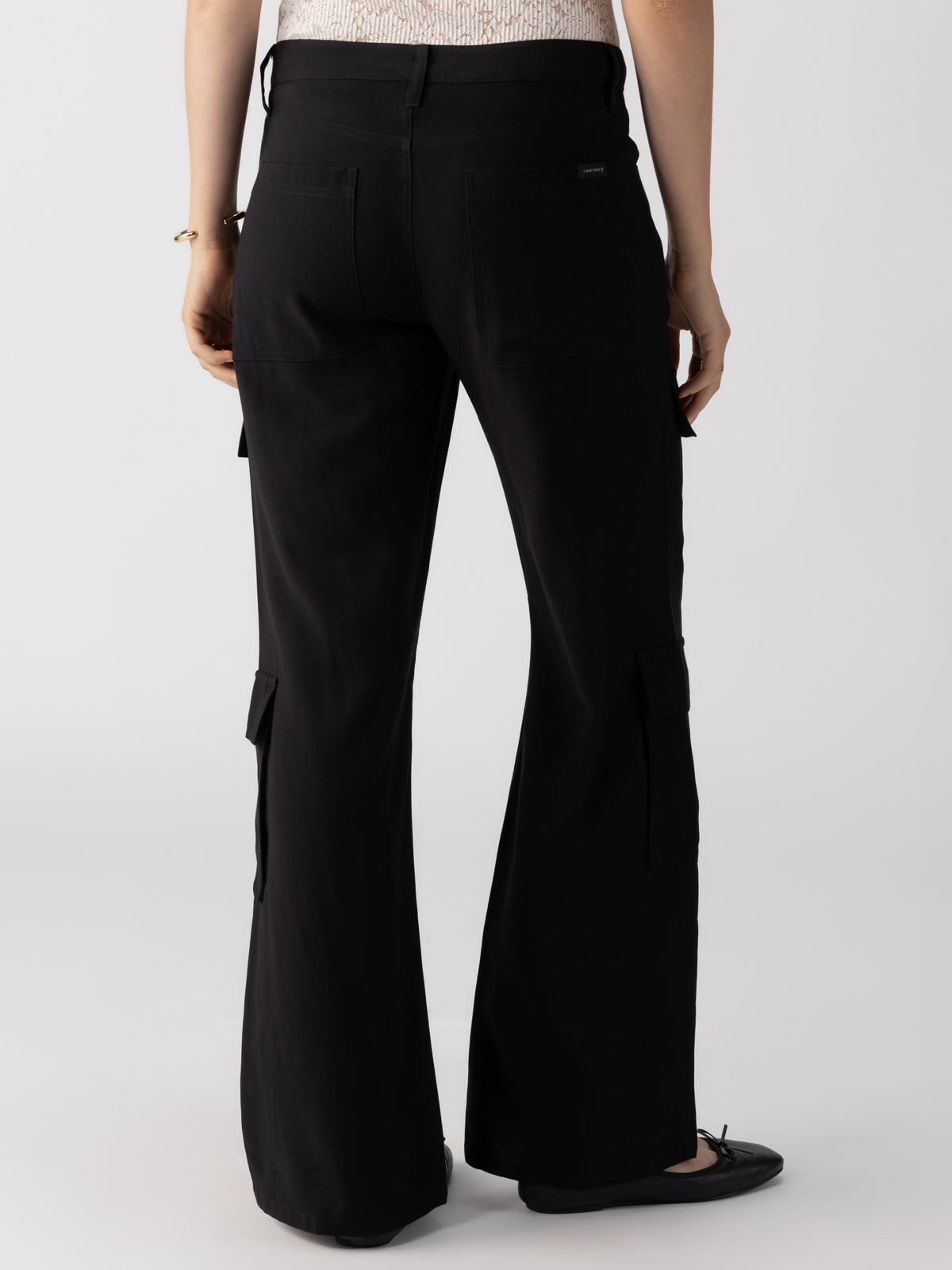 A person is shown from the back, wearing the Low Slung Y2K Pant Black from Sanctuary Clothing, featuring multiple pockets on the legs. They are also wearing a white, sleeveless top and black flat shoes. The background is plain and light gray.