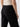 A person is shown from the back, wearing the Low Slung Y2K Pant Black by Sanctuary Clothing, featuring visible side and back pockets. They are also wearing a light-colored, patterned top. The background is plain and neutral.