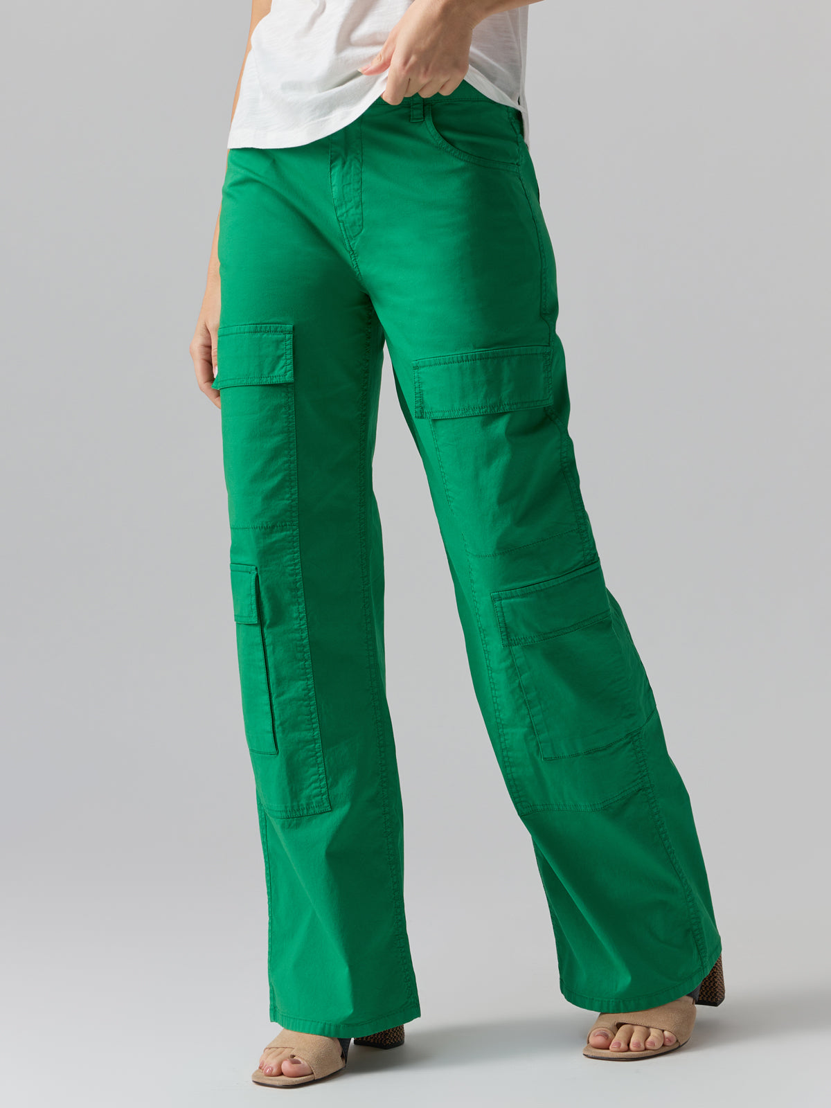 A person is wearing the Sanctuary Clothing 'Low Slung Y2K Pant Jelly Bean' in a bright green shade, paired with a white top, partially visible from the waist down. The pants feature multiple pockets and the person poses with one hand on their hip. They are also wearing open-toe sandals.