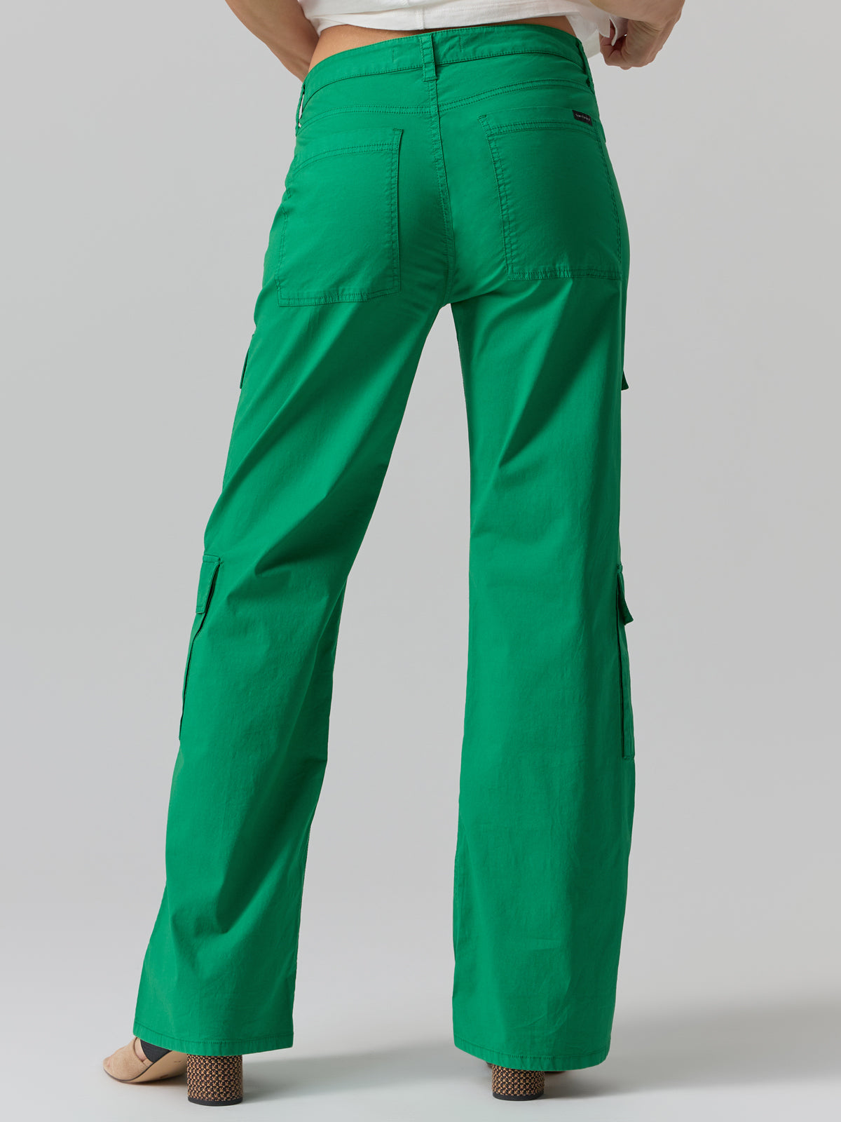 A person is shown from the waist down wearing Sanctuary Clothing's Low Slung Y2K Pant Jelly Bean, featuring large pockets on the back and sides. The loose-fitting, bright green cargo pants are paired with beige open-toe heels, and a portion of a white top is visible at the waist. The background is plain gray.