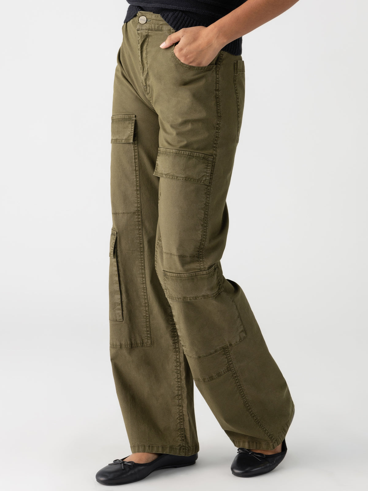 A person is shown from the waist down, wearing the Low Slung Y2K Mossy Green cargo pants from Sanctuary Clothing, featuring multiple pockets and black ballet flats. The pants have a loose fit with a button closure at the waistband against a plain white background.