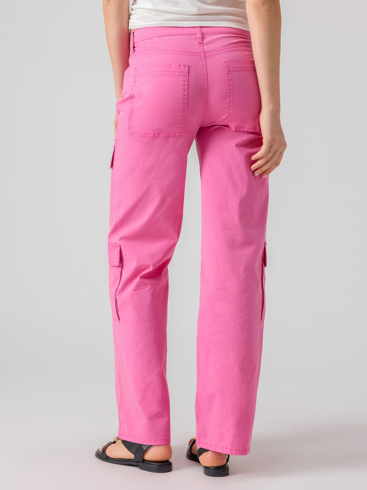 A person wearing Sanctuary Clothing's Low Slung Y2K Wild Pink cargo pants and black sandals is standing with their back to the camera. The pants feature pockets on the back and sides. The background is a plain, light grey color.