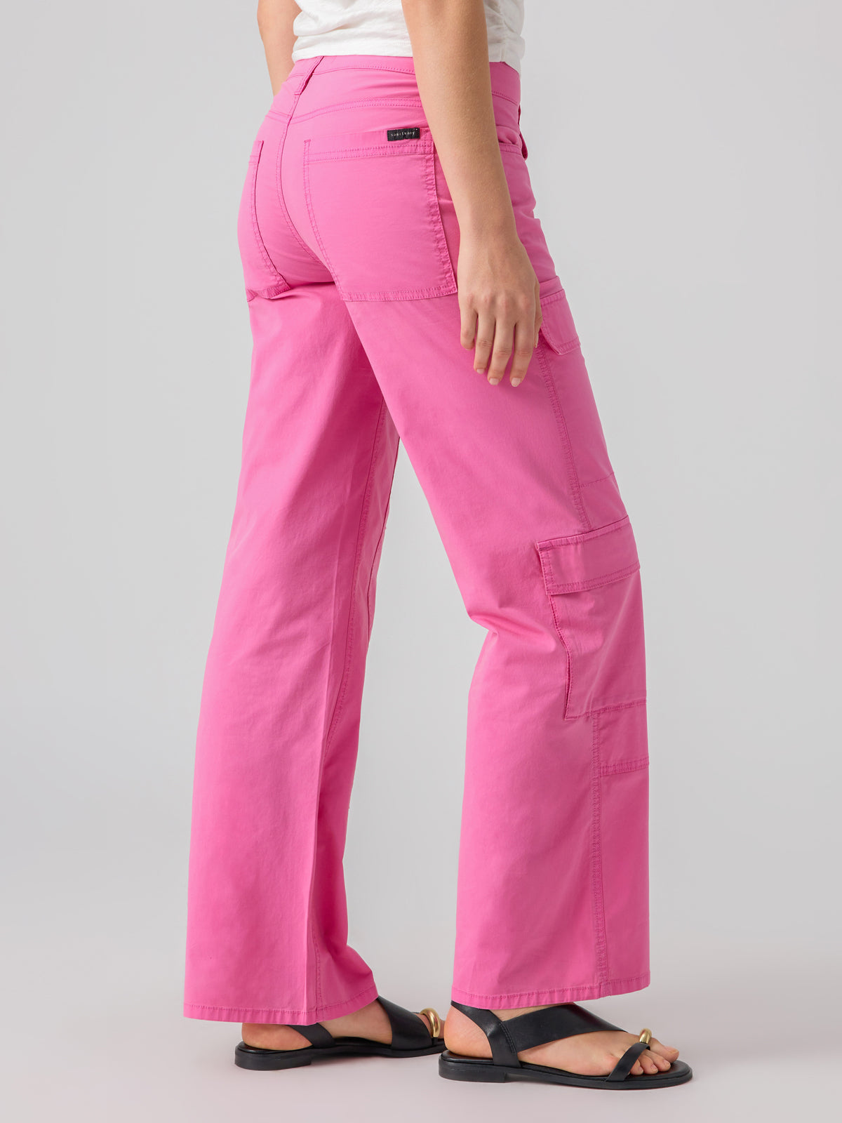 A person is standing sideways, showcasing the Low Slung Y2K Wild Pink cargo pants from Sanctuary Clothing, featuring multiple pockets and a black label on the back pocket. They are wearing a white top and black sandals against a plain light grey background.