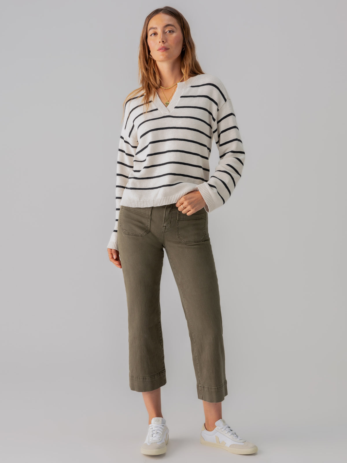 A person with long hair is standing against a plain background, wearing a white and black striped v-neck sweater, The Marine Crop Trouser Pant Fatigue by Sanctuary Clothing in olive green, and white sneakers. They have a relaxed posture with arms at their sides.