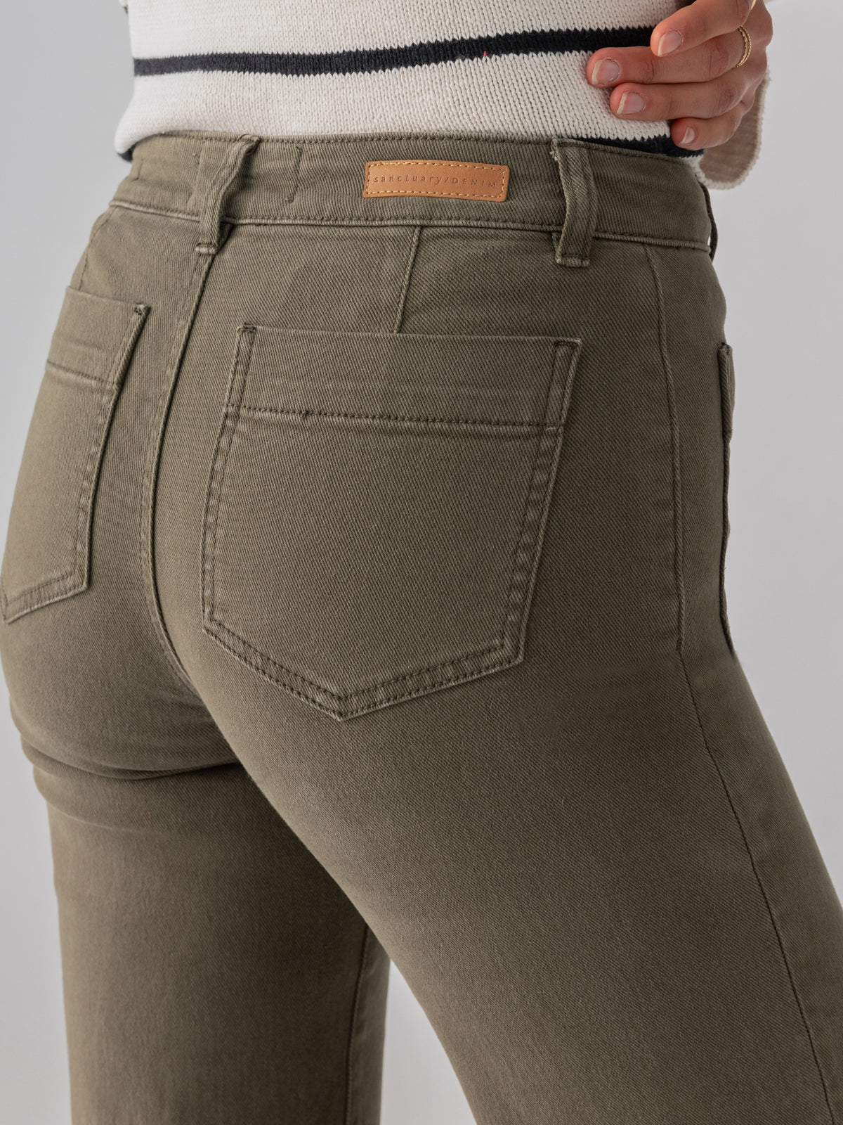 Close-up of a person wearing The Marine Crop Trouser Pant Fatigue by Sanctuary Clothing. The greenish-grey fitted jeans feature visible stitching, belt loops, and back pockets, with a small leather patch on the right side of the waistband. The person is paired with a white and black striped sweater.