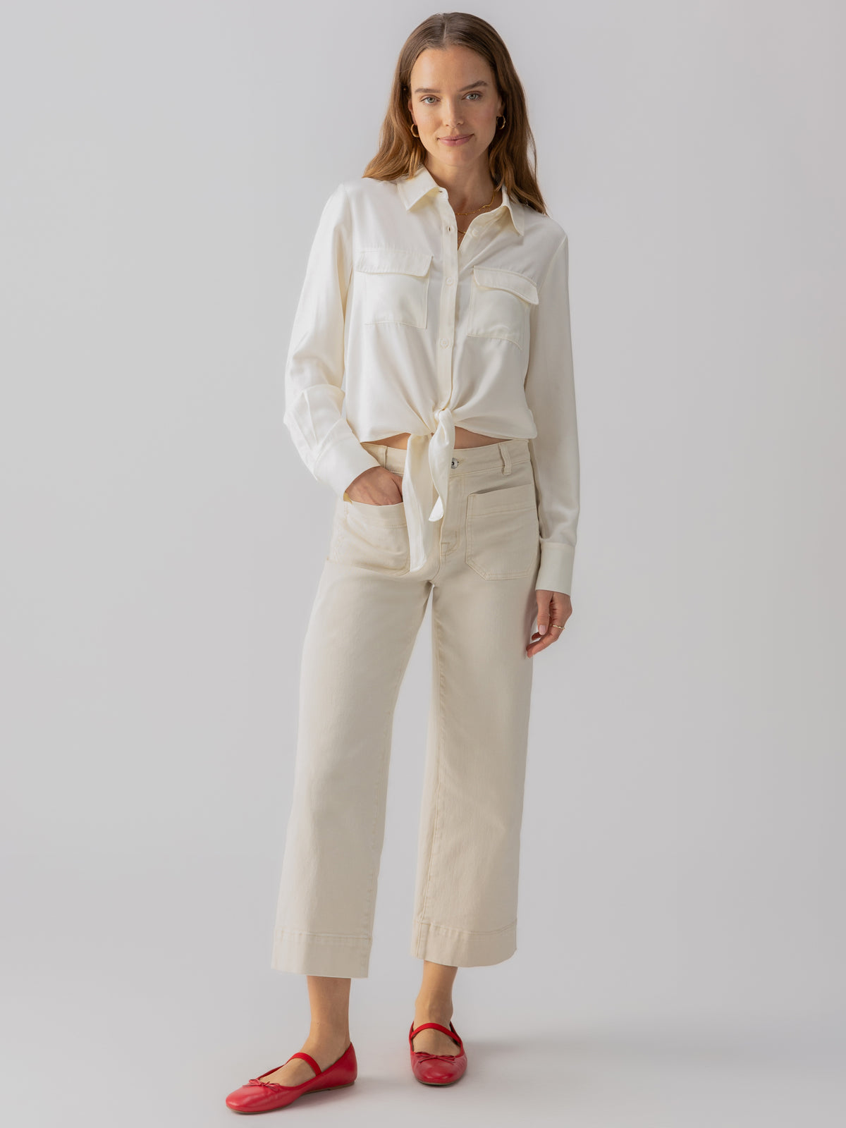 A woman with long hair is wearing a white button-up shirt tied at the waist, The Marine Crop Trouser Pant in French Vanilla by Sanctuary Clothing, and red flat shoes. She stands against a plain light-gray background, looking at the camera.