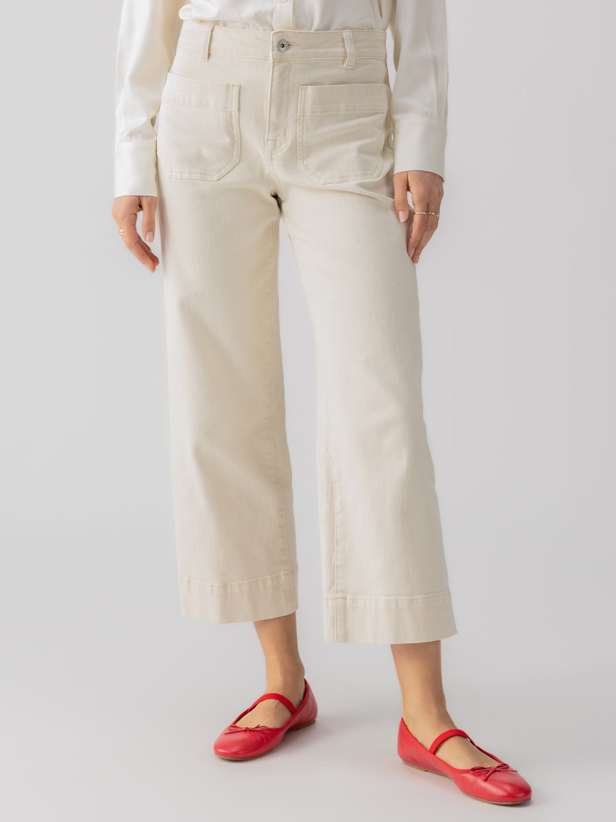 A person is standing against a plain background, wearing The Marine Crop Trouser Pant in French Vanilla by Sanctuary Clothing. They pair the cream-colored high-waisted, wide-leg pants with a white long-sleeve top. Their feet are adorned with red ballet flats, and their hands display a few rings. The overall look is minimalist and chic.