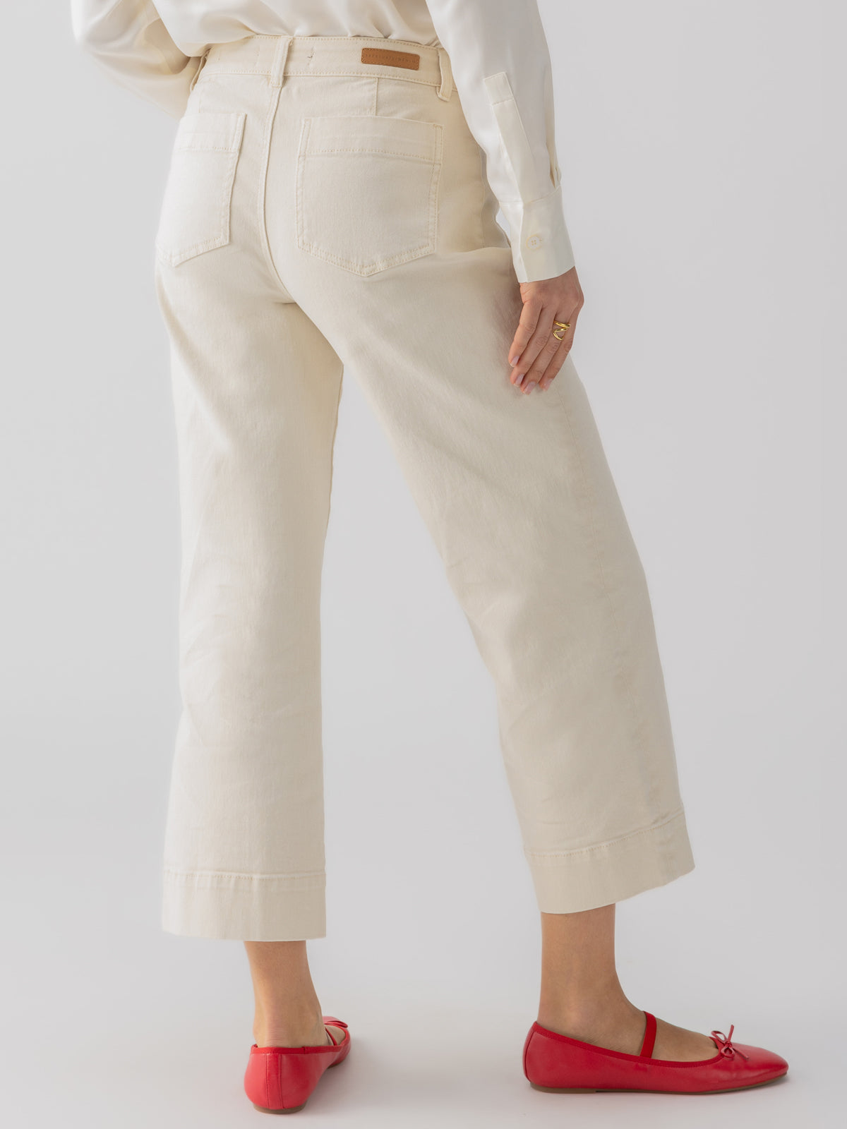 A person is standing with their back to the camera, wearing The Marine Crop Trouser Pant in French Vanilla by Sanctuary Clothing, paired with a white shirt and red ballet flats. The person's left hand is partially visible, resting at their side with a ring on their finger.