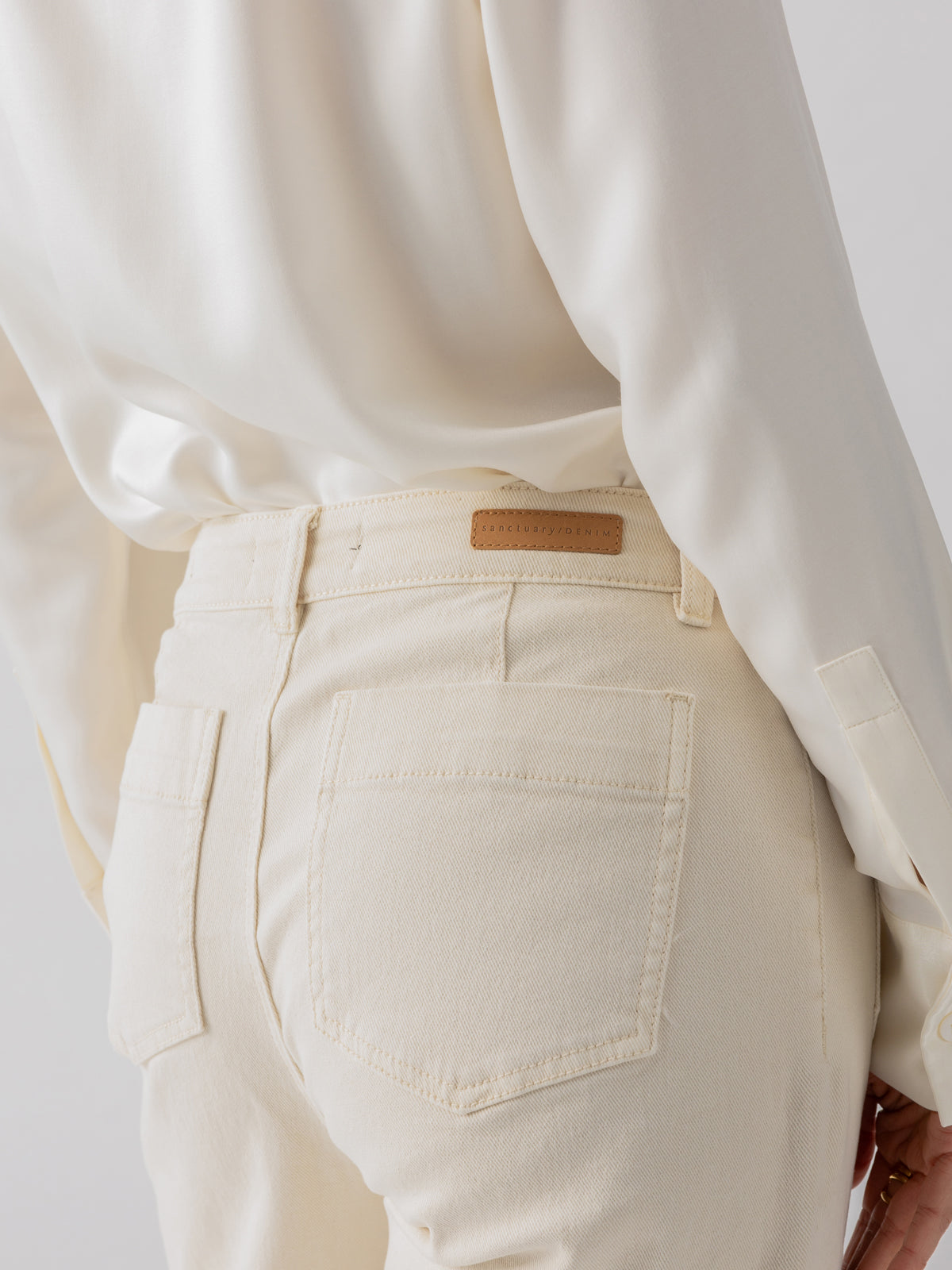 A person is wearing The Marine Crop Trouser Pant in French Vanilla by Sanctuary Clothing, featuring visible back pockets and a tan leather patch near the waistband. They are also wearing a cream long-sleeved shirt. The background is plain and light-colored.