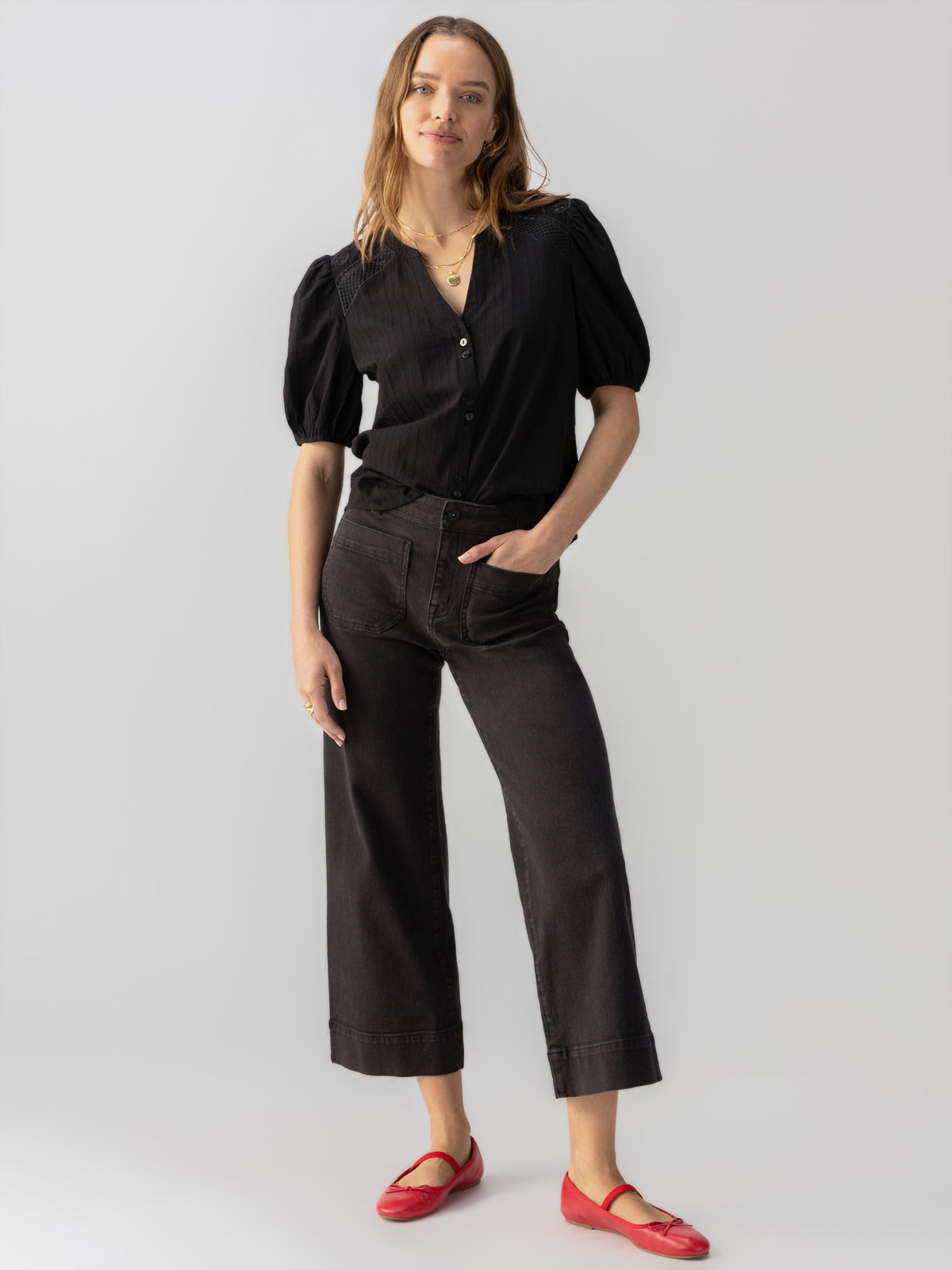 A person standing against a plain background, wearing the Lift Me Up Blouse Black from Sanctuary Clothing with short puffed sleeves, black wide-legged pants, and red flats. They have long hair and one hand in their pocket. They are looking directly at the camera with a relaxed posture.