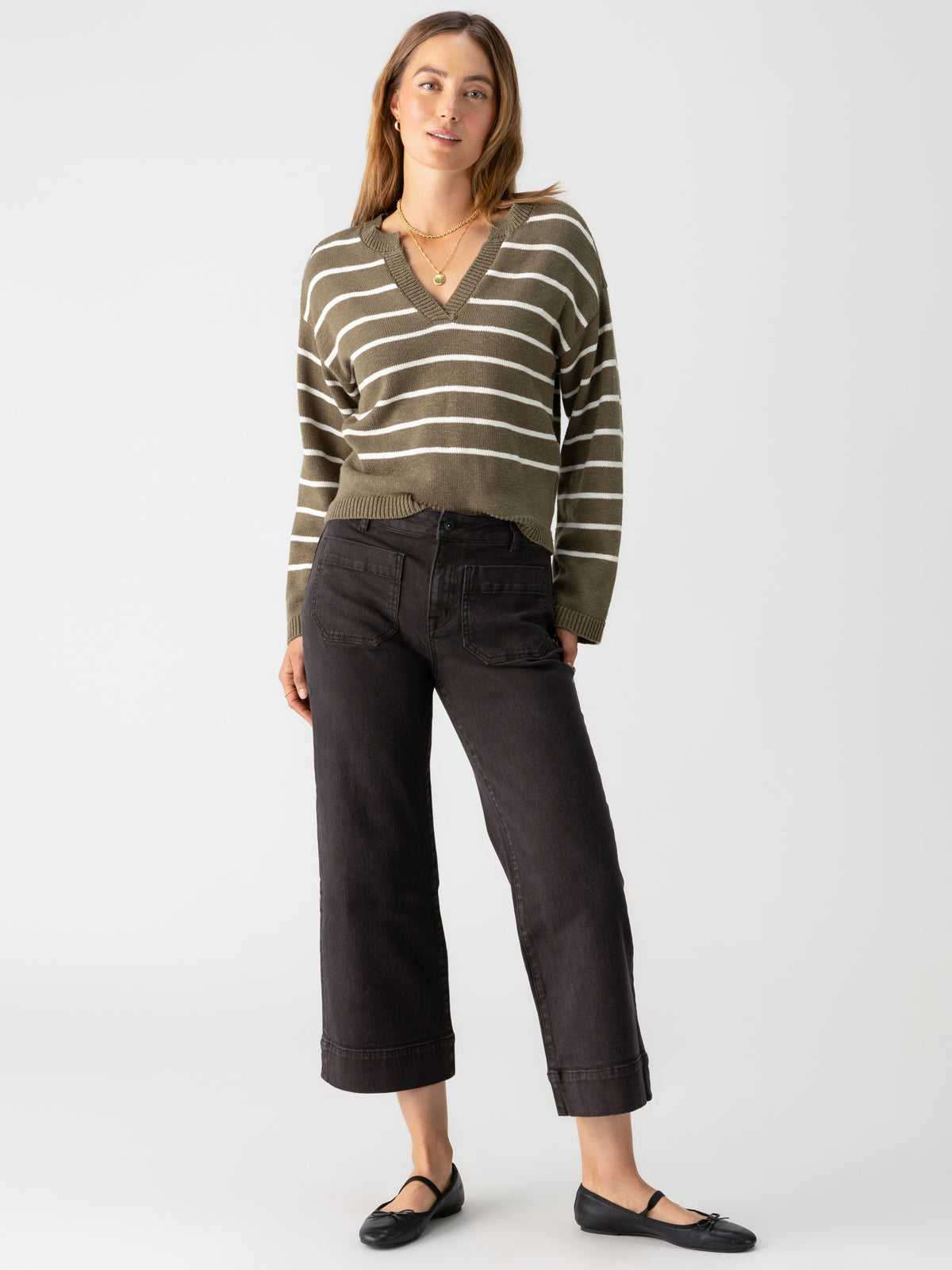 A woman stands against a plain background wearing The Marine Obsidian by Sanctuary Clothing, a green and white striped V-neck sweater, high-waisted black wide-leg jeans, black ballet flats, and a gold necklace. She has long, light brown hair and a composed expression.