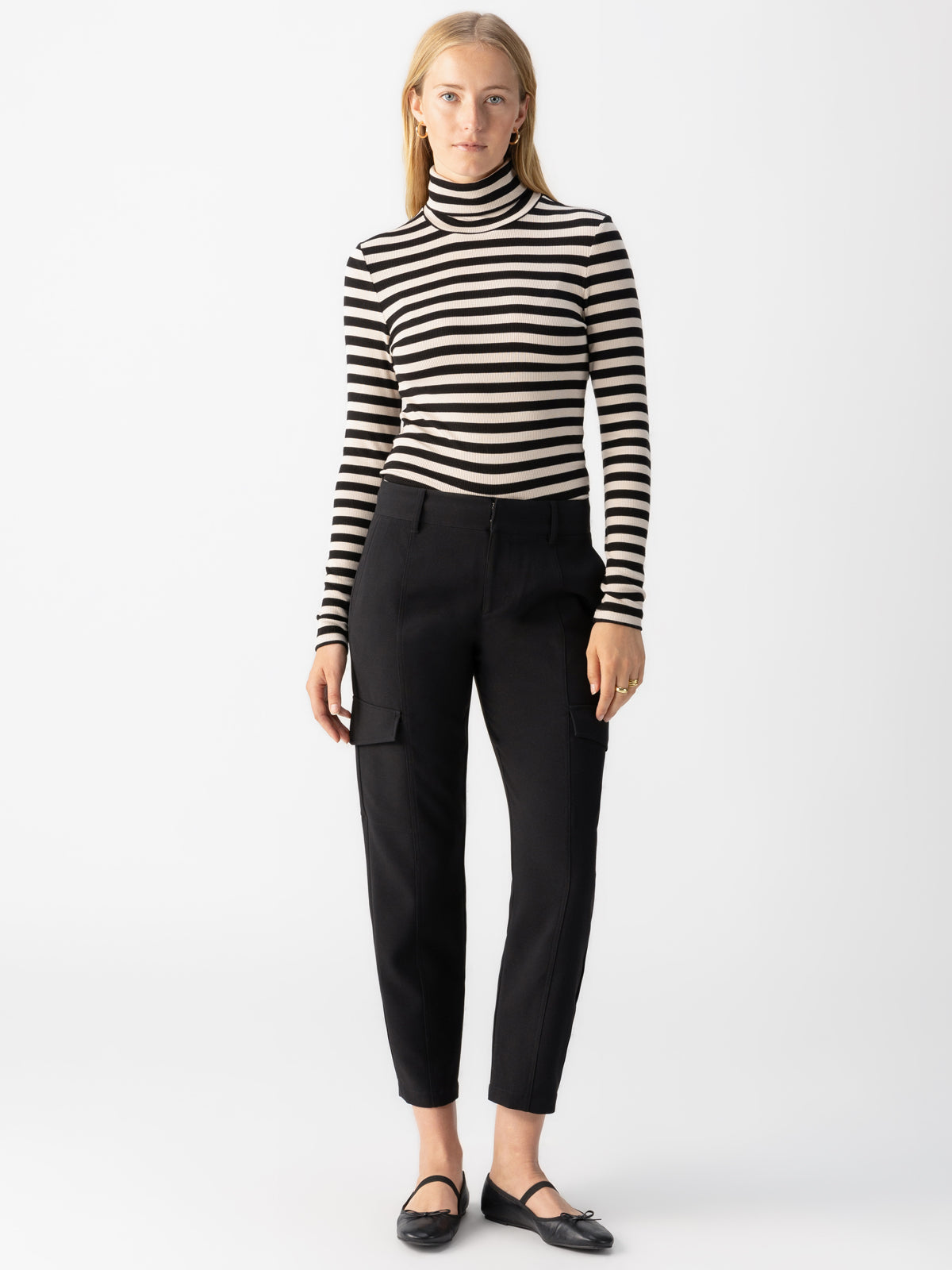 A person with long blonde hair is standing against a plain white background, wearing a black and white striped turtleneck sweater, Polished Cargo Pant Black by Sanctuary Clothing, and black ballet flats. They have a neutral expression and arms hanging by their sides.