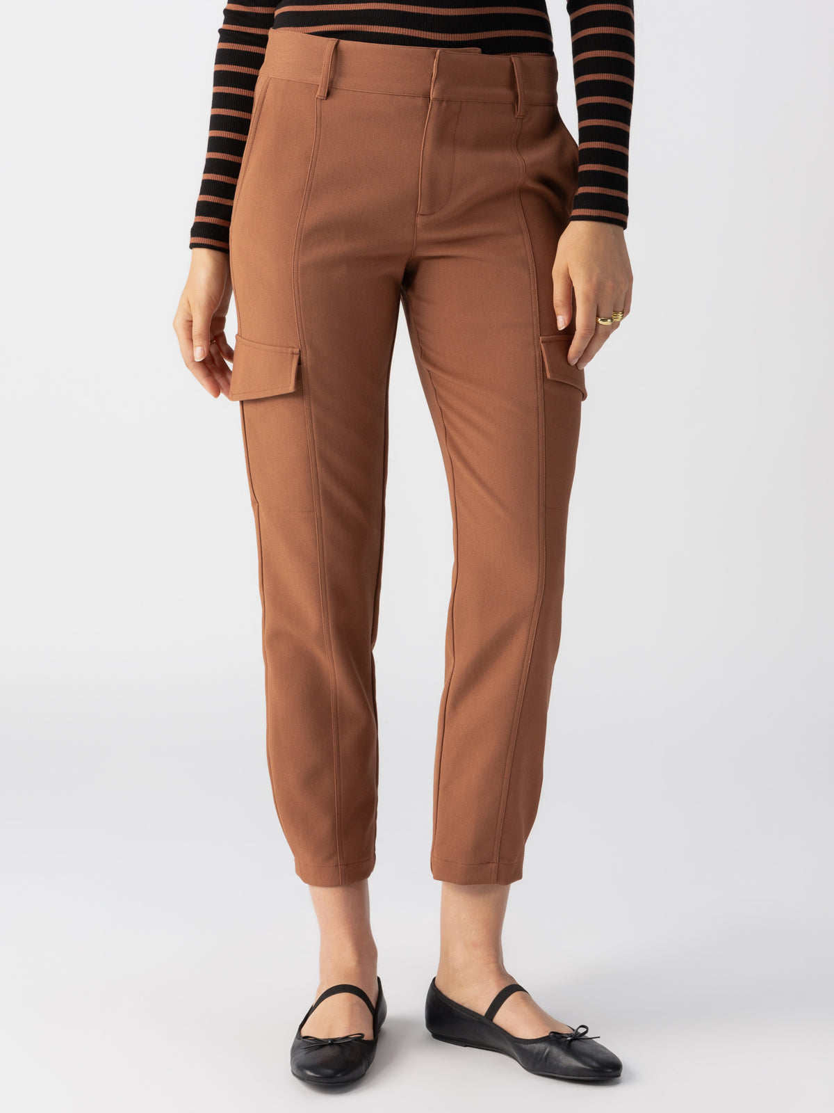 A person is wearing Sanctuary Clothing's Polished Cargo Raw Hide pants, which are brown with large side pockets, paired with a black and brown striped long-sleeve top. They are also wearing black ballet flats. The background is plain and white, drawing attention to the outfit.