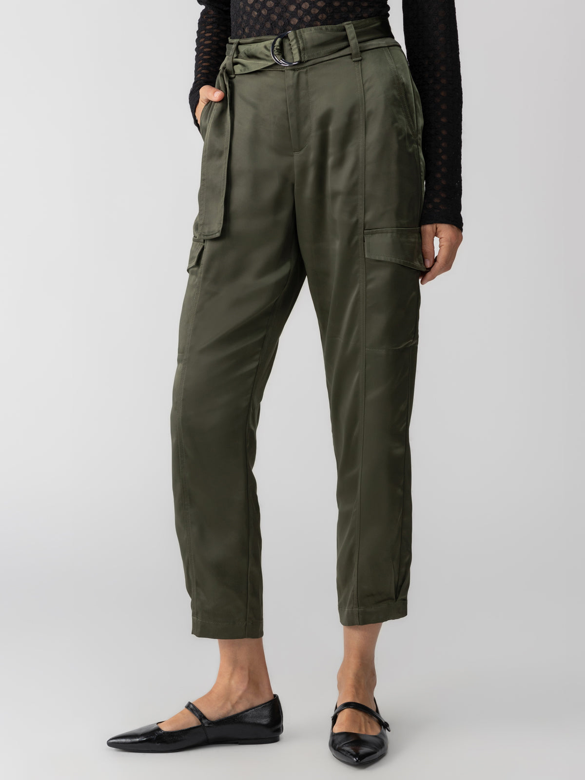 A person wearing Sanctuary Clothing's Classy Cargo Trouser in Dark Olive, paired with a black belt and a black long-sleeve top. The trousers feature multiple pockets and are matched with flat, closed-toe black shoes against a plain light gray background.