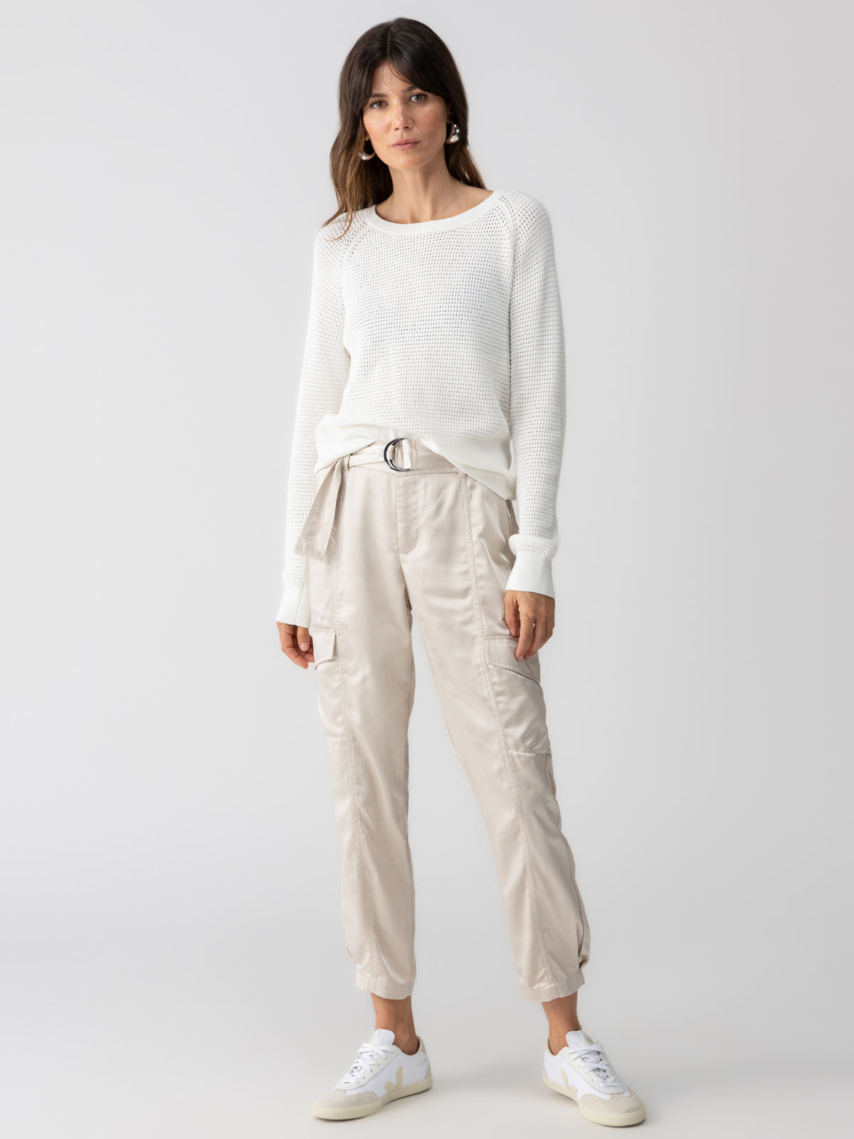 A woman stands against a plain background wearing a white knit sweater and the Classy Cargo Trouser Pant Toasted Marshmallow by Sanctuary Clothing. She has long brown hair and is wearing white sneakers. Her hands rest at her sides, and she looks directly at the camera with a neutral expression.