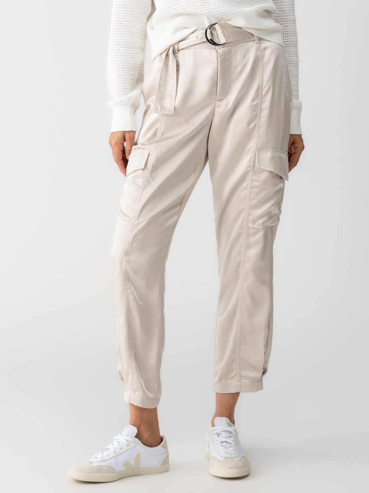 A person wearing the Classy Cargo Trouser Pant Toasted Marshmallow by Sanctuary Clothing, along with a belt and multiple pockets, stands with their hands by their sides. They are also dressed in a white sweater and white sneakers. The background is plain and white.