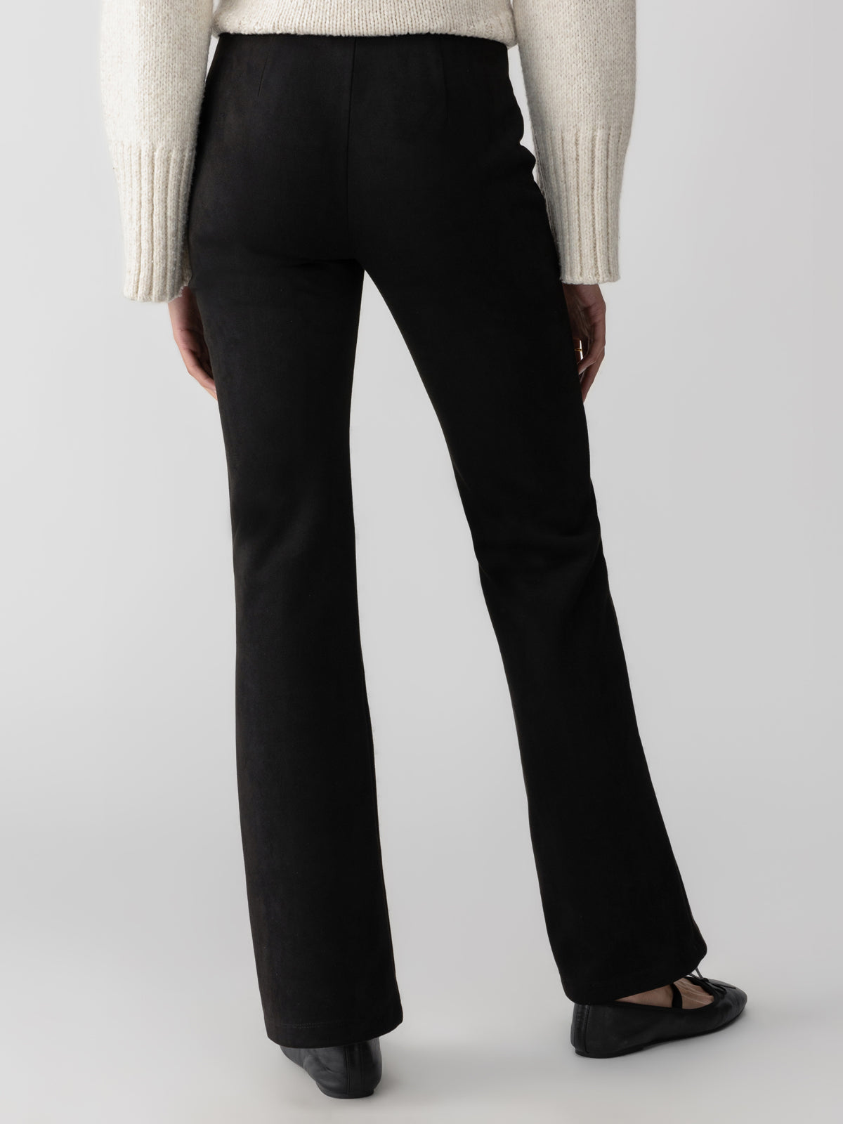 A person wearing a white, long-sleeved sweater and Sanctuary Clothing's Lana Flare Black pants stands facing away, highlighting the back of the outfit. The person is also wearing black shoes, set against a neutral gray background.