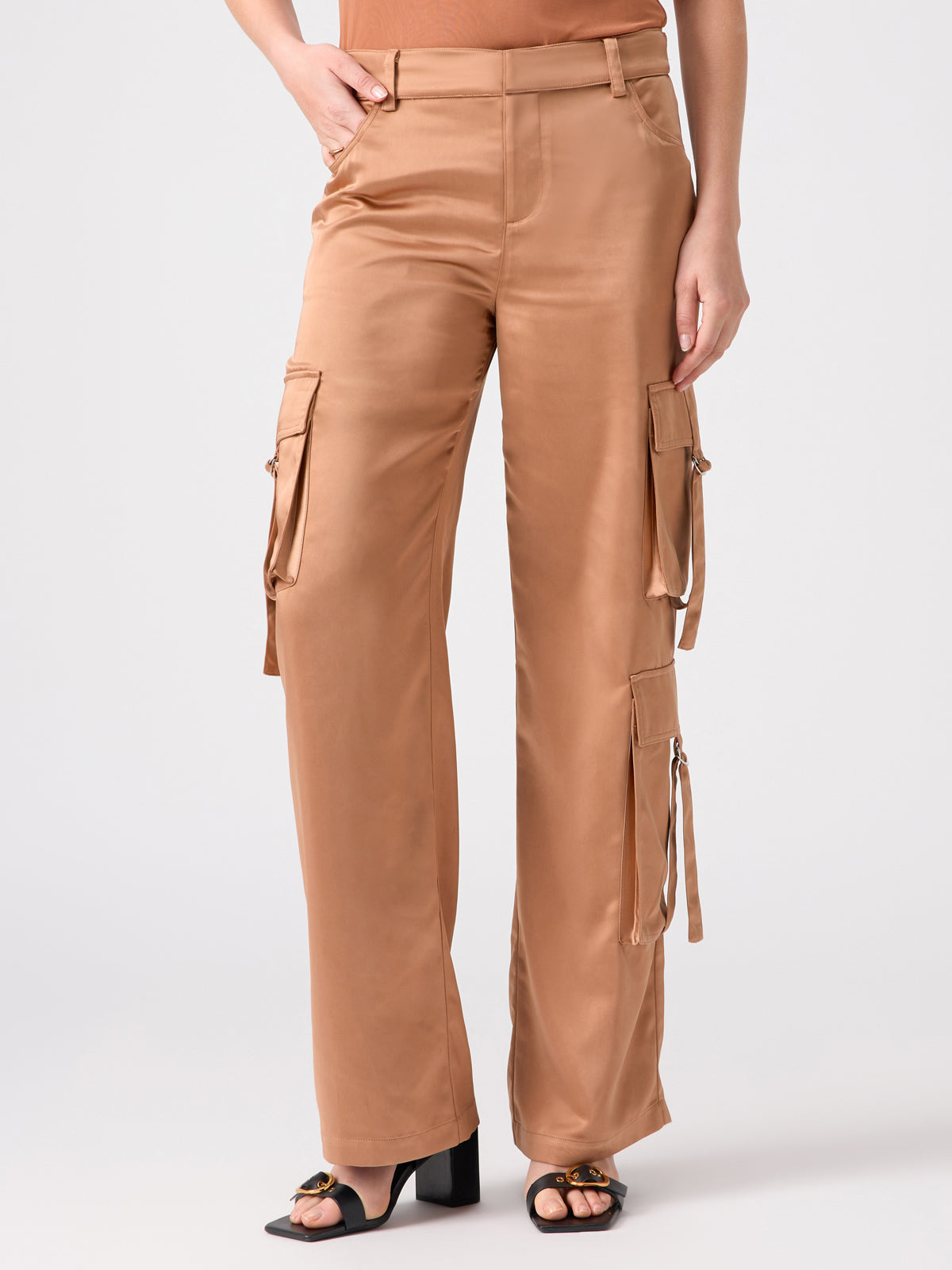 A person wearing the Y2K Strappy Cargo Mocha Mousse pants from Sanctuary Clothing, featuring a high-waisted, wide-leg design in tan with multiple pockets and side buckles, paired with open-toe block-heeled sandals. The background is plain white.