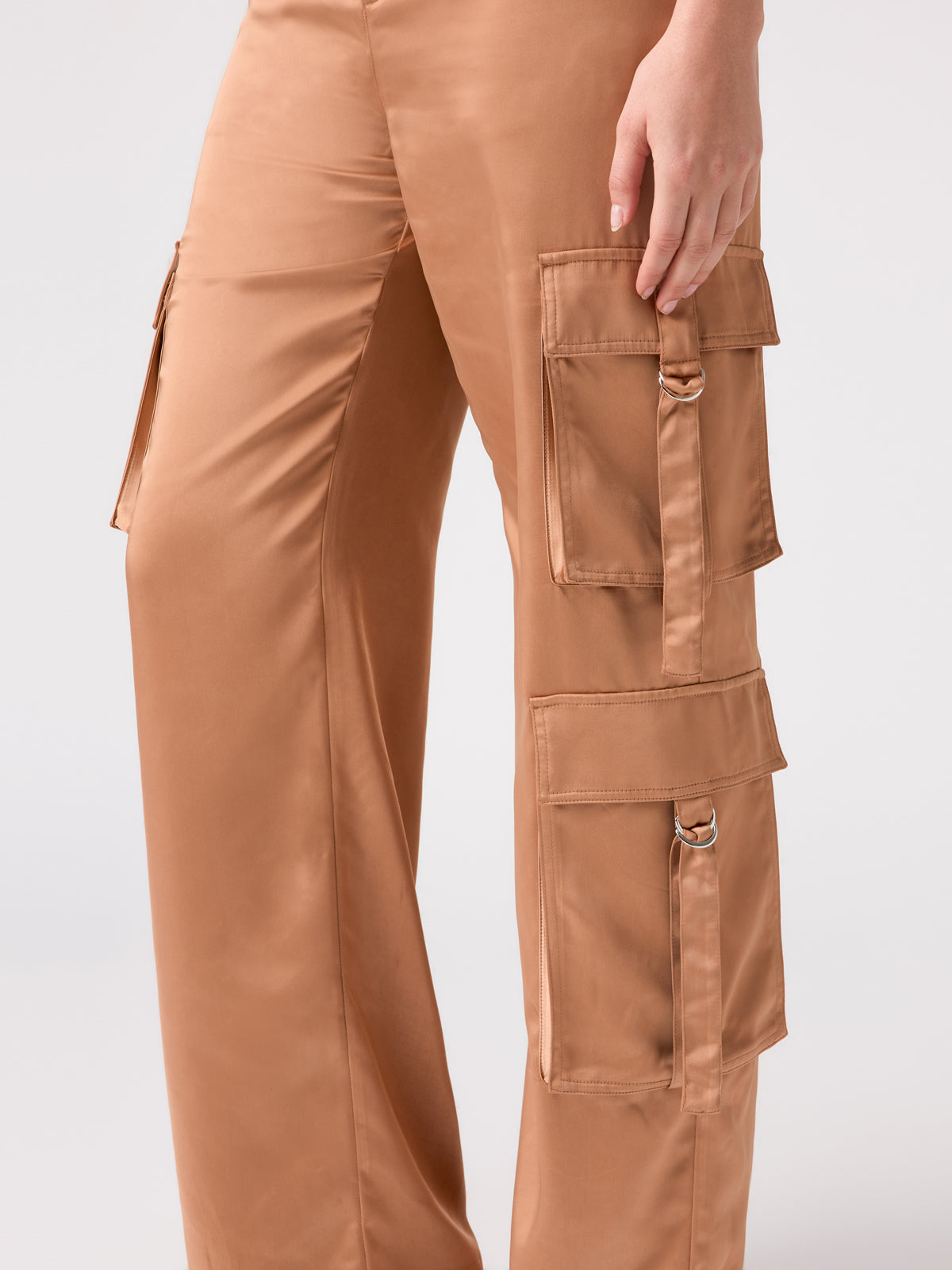 A person is wearing the Y2K Strappy Cargo Mocha Mousse pants from Sanctuary Clothing. The satin fabric features multiple pockets with flaps and metal D-ring accents. The person's hand is resting on the right thigh, set against a plain, light-colored background.